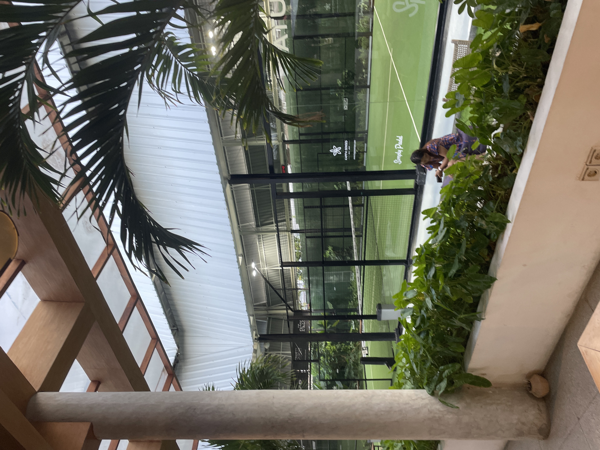 The image depicts a scene at a paddle tennis court in Denpasar, Indonesia. The court, visible through a glass enclosure, has a green playing surface. A person is standing near the court's edge, possibly observing or interacting with the game. Lush green plants are positioned along a wall, bordering the court area. The architecture features wooden beams and a white roof structure.