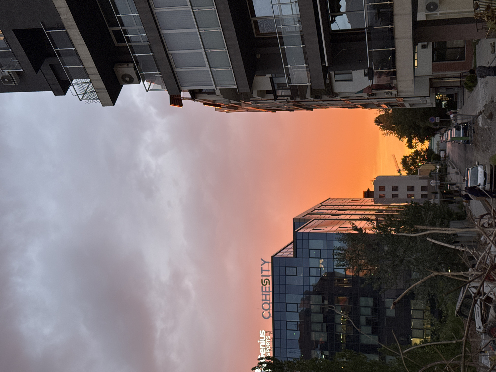 The image captures a street scene in Sofia, Bulgaria, featuring buildings under an orange sky at sunset or sunrise. The view is looking up between two opposing rows of buildings. On the left, there are multi-story residential buildings with balconies and air conditioning units, facing skyward. On the right, there is a modern glass office building with a sign reading "Cohesity" and "Genius Sports" going up the building, indicating commercial activity. The street below includes parked cars and people walking, suggesting an active urban environment. A construction crane can be seen further down the street. The sky transitions from a grey, overcast color on one side to a vivid orange in the middle, indicating a sunrise or sunset.