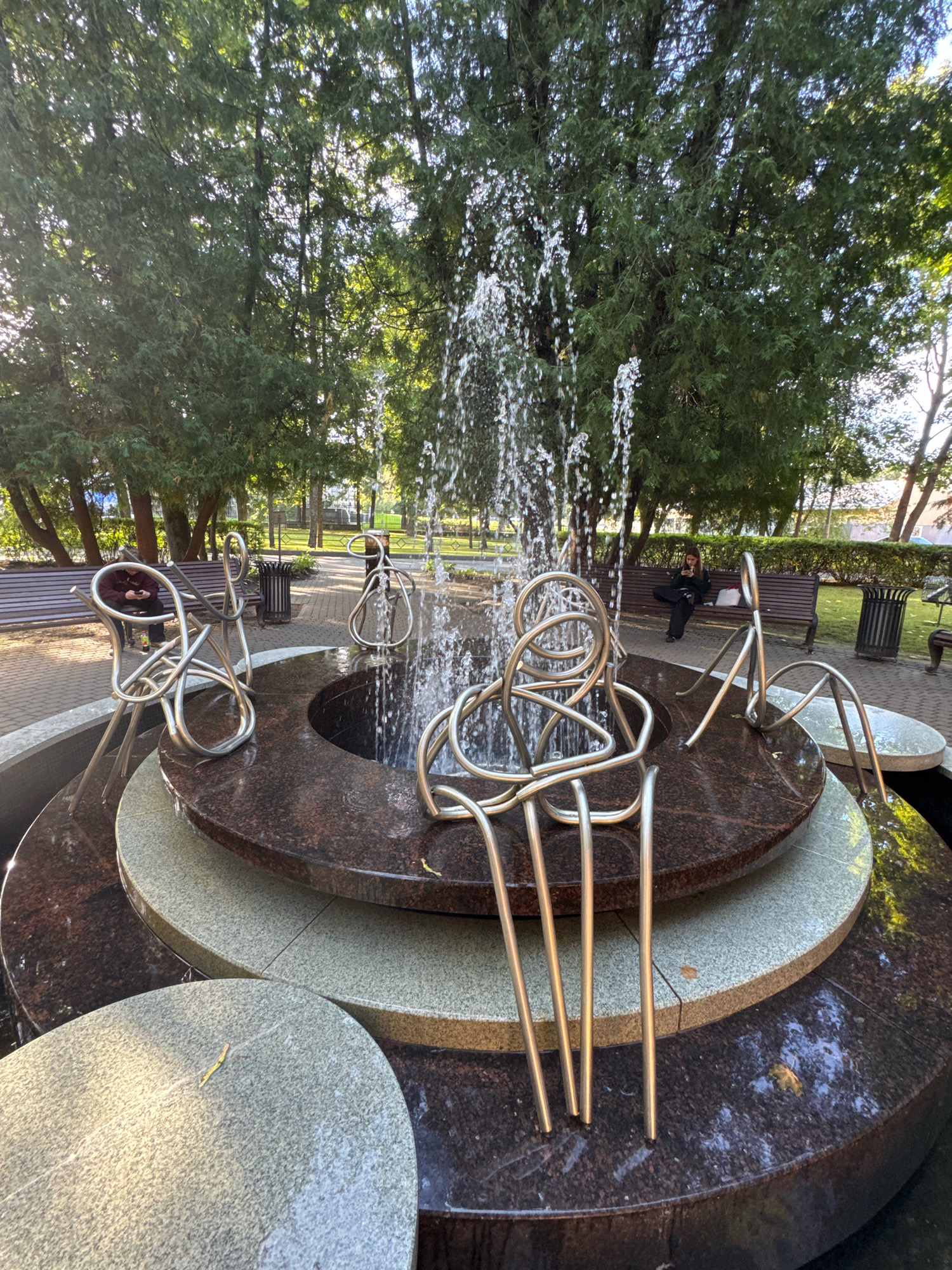 The image shows a multi-tiered fountain with abstract metal sculptures in the foreground, located in a park in Saint Petersburg, Russia. The fountain is surrounded by granite tiers, with water spraying upwards in the center. Behind the fountain, several people are visible sitting on park benches and walking. The environment is lush with green trees and foliage, and the ground is paved with cobblestones. The overall atmosphere is peaceful and serene, with the sunlight filtering through the trees.