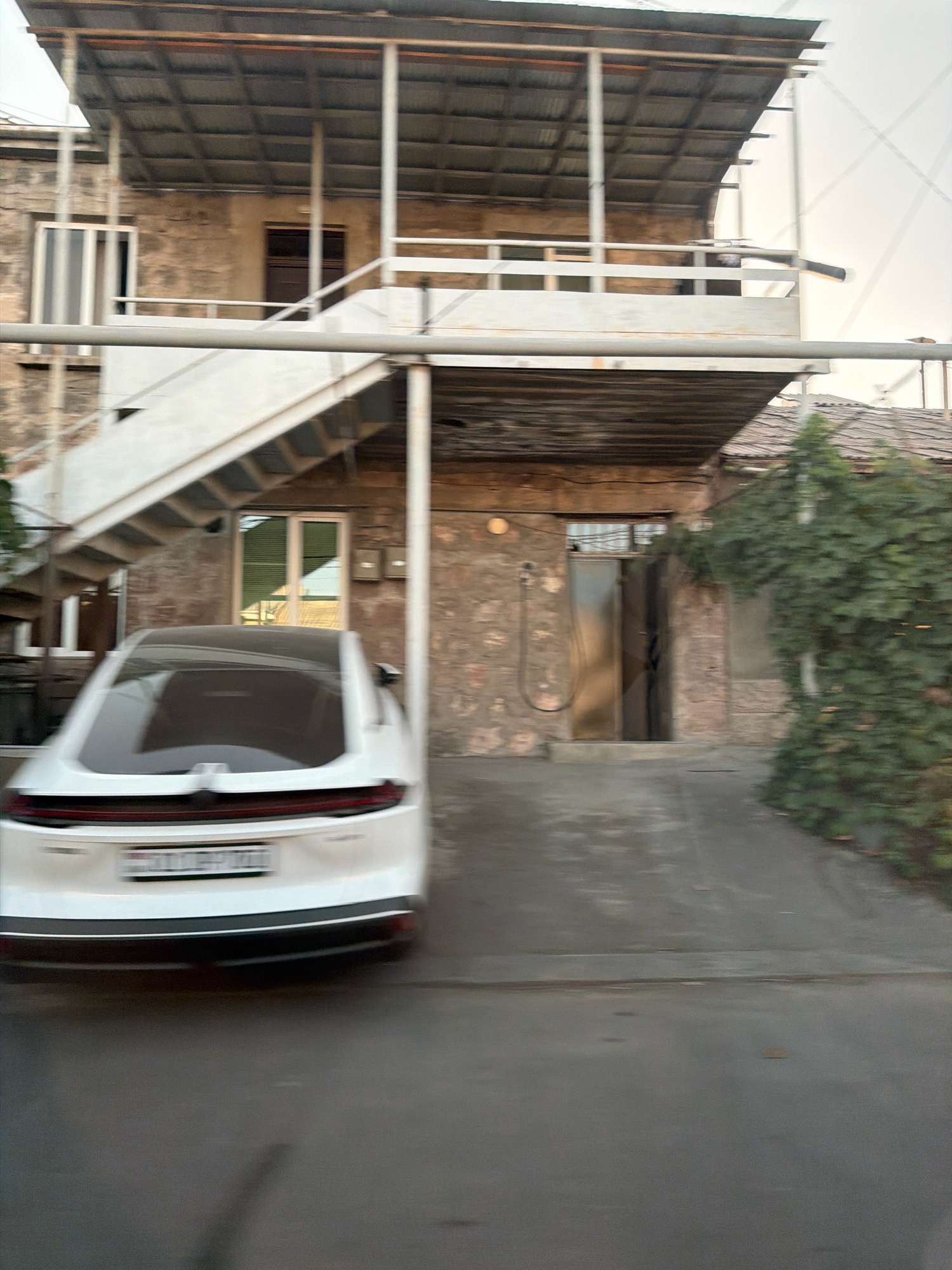 The image captures a street scene in Yerevan, Armenia, focusing on a white car parked in front of a two-story building with an external staircase. The building appears to be constructed of stone and has a corrugated metal roof. The car, a modern sedan, is partially blurred due to motion, suggesting it was either captured while moving or the photograph was taken from a moving vehicle. The license plate of the car is visible but illegible due to motion blur. The environment suggests a residential or mixed-use area within the city, with some greenery visible to the right of the building. The overall impression is a candid snapshot of urban life in Yerevan.