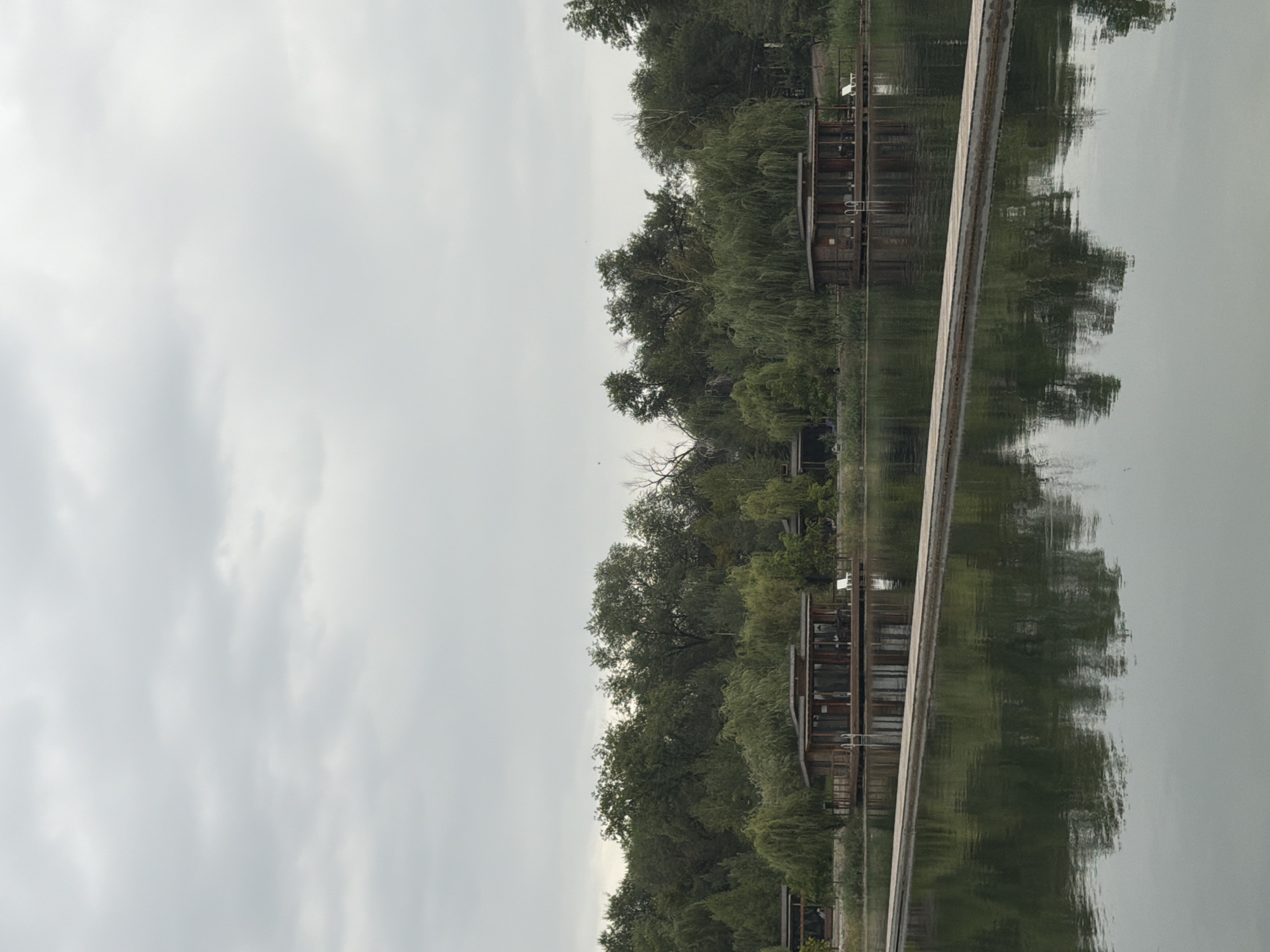 The image depicts a serene lakeside scene in Pervomayskiye Prudy, Kazakhstan. The foreground is dominated by a calm body of water reflecting a cloudy, overcast sky. A wooden walkway extends across the water, creating a clear division between the foreground and midground. In the midground, several modern, wooden structures with large glass windows are situated along the shoreline, appearing to be waterfront cabins or bungalows. These structures are surrounded by lush green trees and vegetation, forming a dense backdrop. The overall atmosphere is tranquil and natural, suggesting a resort or a peaceful residential area. There are no people visible in the image.