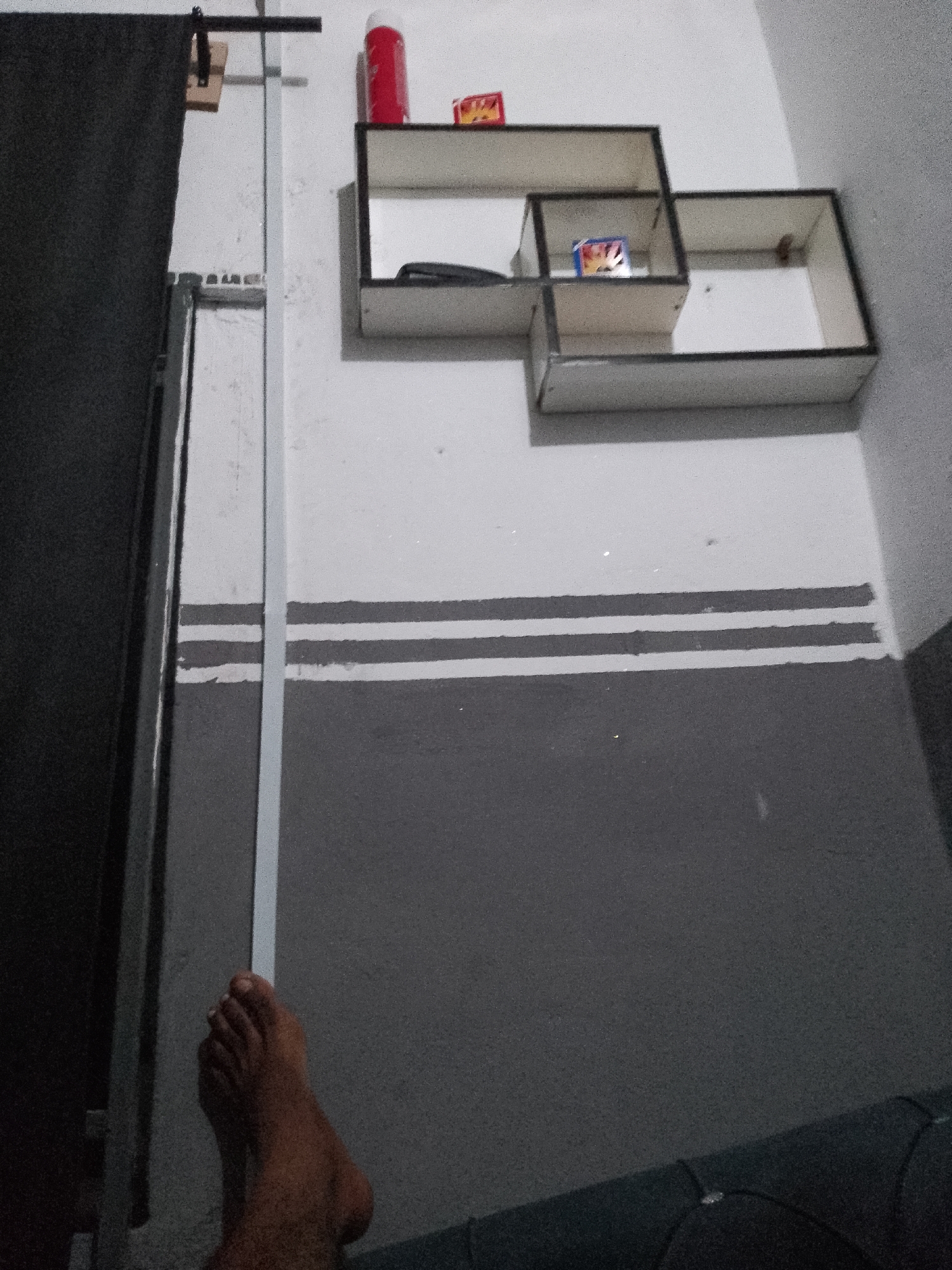 A human foot, with dark skin and clearly defined toes, rests on a dark surface, possibly a sofa, in the foreground. Behind the foot, a tall, thin, grey vertical support runs down the center of the image. To the left of the support, a dark, textured material, likely a curtain or drape, hangs down. The background wall is painted in two horizontal sections: the upper section is light grey, and the lower section is a darker grey. Between these two sections, there are three distinct horizontal stripes, the top and bottom stripes are dark grey, and the middle stripe is light grey.
Mounted on the upper, lighter grey section of the wall are two overlapping, white, cube-shaped shelves with black trim. The left shelf, positioned higher, contains a red aerosol can and a small, brightly colored rectangular object, possibly a pack of cards or candy. The right shelf, positioned lower and slightly to the right, is empty except for a small, brown, circular object. A black object, possibly a phone, is visible inside the left shelf, partially obscured.
To the far left of the image, near the top, a wooden block is attached to a black horizontal bar, which is itself attached to the vertical support. A thin, light grey vertical bar runs alongside the dark curtain. The overall lighting is dim, creating shadows and a somewhat grainy appearance to the image. The scene appears to be an indoor domestic setting.