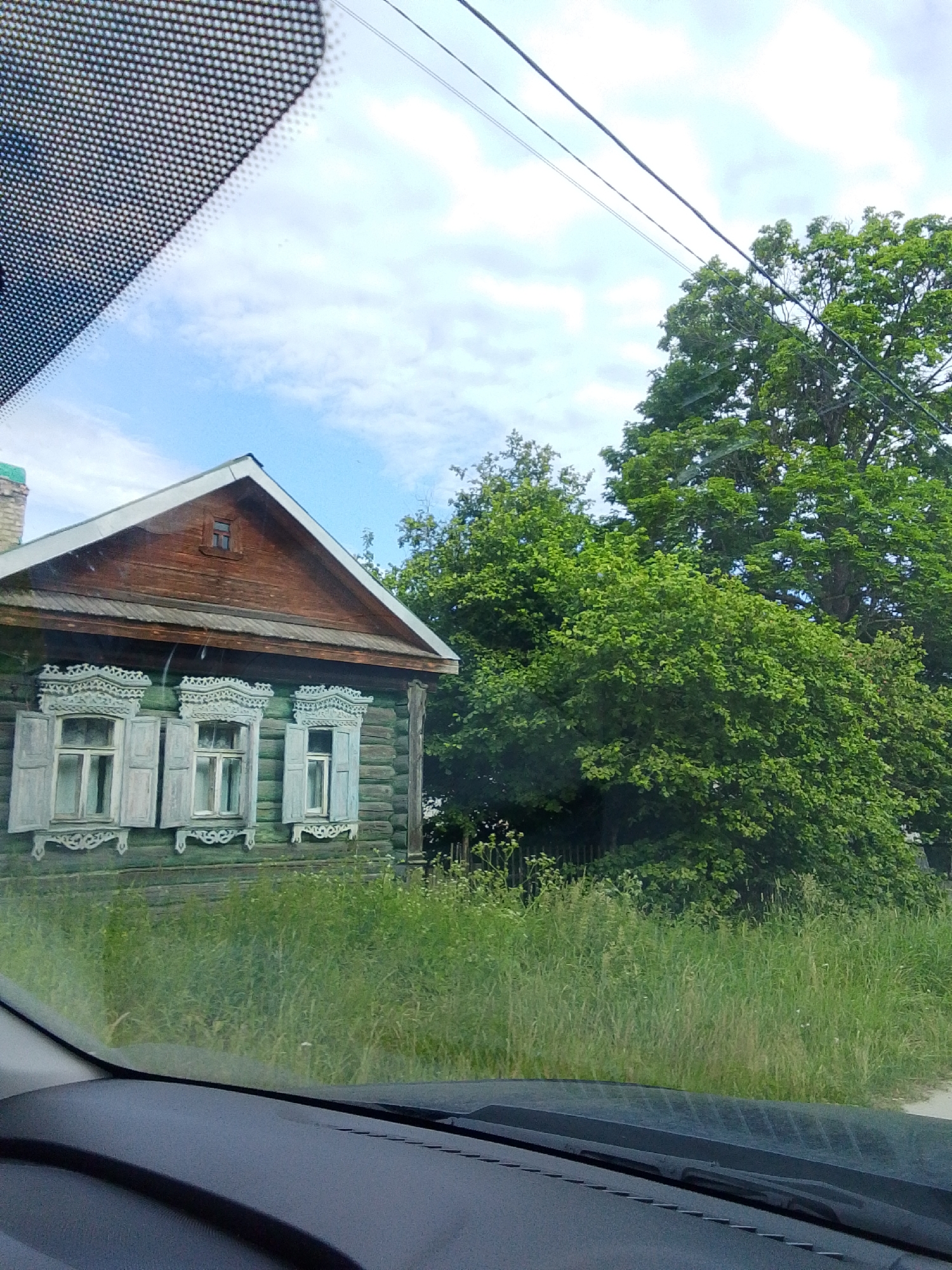 This media file captures a view from inside a vehicle, looking out at a traditional wooden house in Staryi Pokrov, Russia. The house, with its green wooden walls and ornate white window shutters and trim, appears to be quite old. The roof is made of dark wood, and a small window is visible on the gable.
The house is surrounded by tall, green grass and lush trees, suggesting a rural or suburban setting in a temperate climate. The sky is partly cloudy, with patches of blue visible, indicating daytime and fair weather. A power line runs diagonally across the upper portion of the frame.
The perspective suggests the viewer is looking out through a windshield, with a portion of the car's dashboard visible in the foreground. There are no people visible in the image, and the overall impression is one of a quiet, picturesque rural landscape. The time of day appears to be late morning or early afternoon. No text is visible in the image.