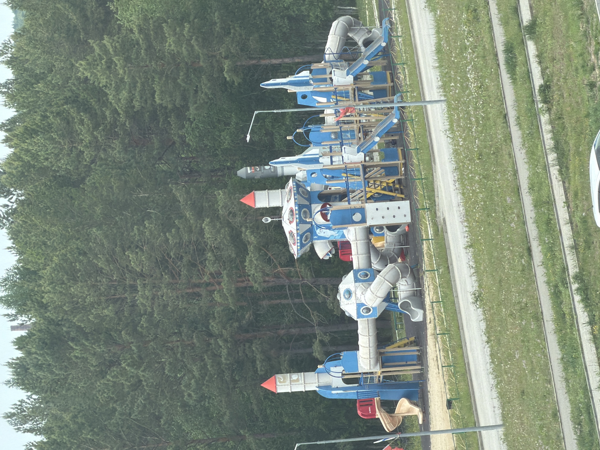 A large, multi-component playground designed with a space theme is situated in an outdoor setting with a dense forest in the background. The playground features multiple rocket-shaped structures, interconnected tubes, slides, and climbing elements, predominantly in white and blue colors with red accents. The ground beneath the playground appears to be covered with sand and wood chips. A paved road runs in front of the playground, separated by a grassy area with sparse white wildflowers and a low green fence. Several lampposts are visible along the road. The overall environment suggests a park or recreational area bordering a forested region, likely within the city of Tsiolkovsky, Russia, given the thematic elements relating to space exploration and the presence of coniferous trees characteristic of the region. No people are visible in the scene.