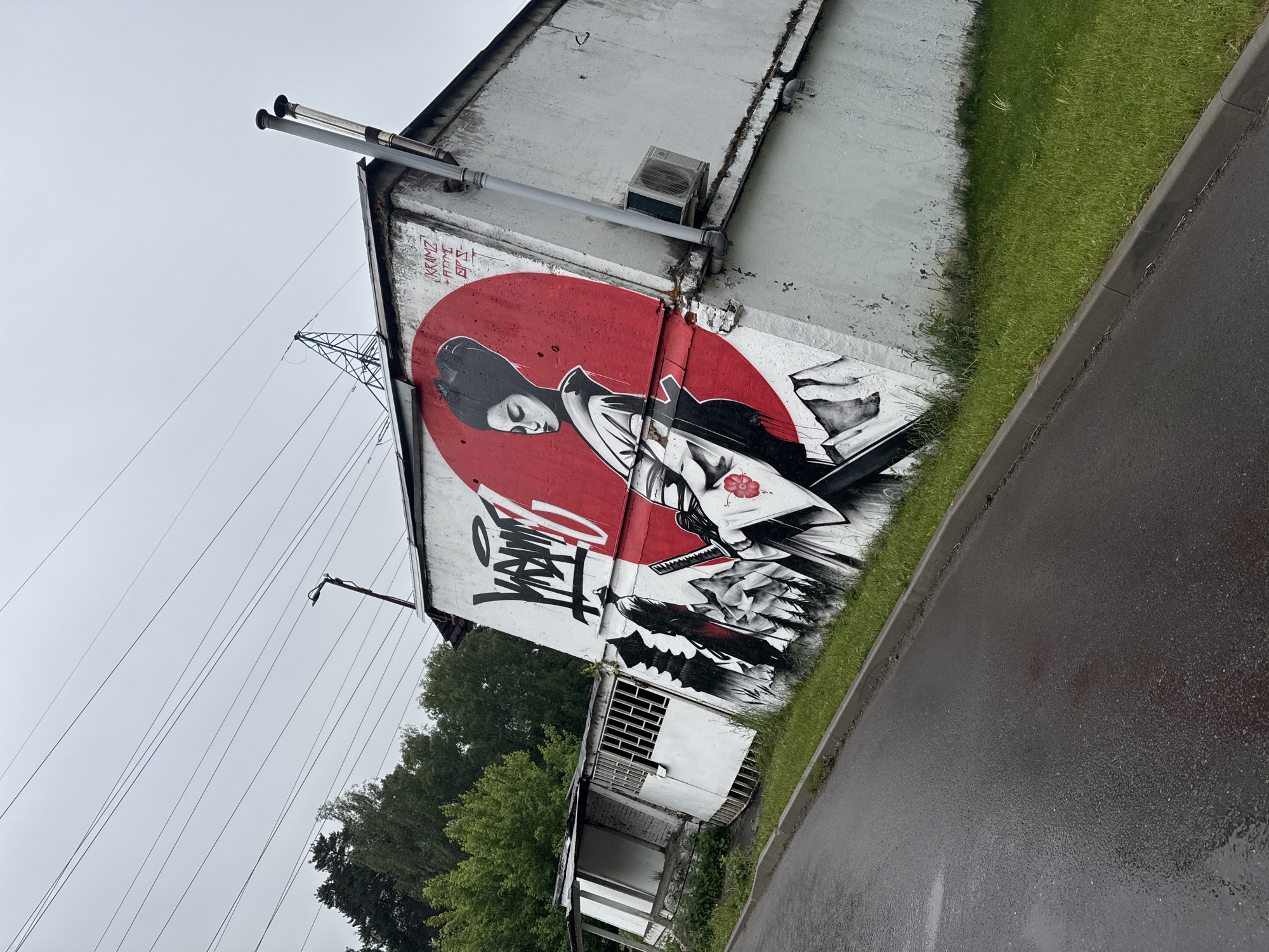The image shows a building exterior in Balashikha, Russia, featuring a large mural depicting a geisha against a red circle, with the "Stussy" logo incorporated into the design. The mural is painted on a light gray, slightly weathered building facade. Above the mural, there are pipes and an air conditioning unit attached to the building. Below the mural, there is a partially enclosed, small structure attached to the building, with visible ventilation grids and some vegetation growing around it. A patch of grass separates the building from a wet, asphalt road. Power lines and poles are visible in the background against a cloudy sky. Trees are present in the background.
