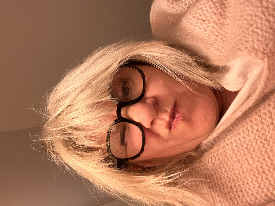 A woman with blonde hair and glasses looks down at the camera. She wears a light pink, knitted sweater with a hood, which is drawn up around her face. Her expression is slightly pensive, with lips pursed. Light illuminates one side of her face. The background includes a beige or light gray wall. The image is rotated, causing the subject to appear sideways.