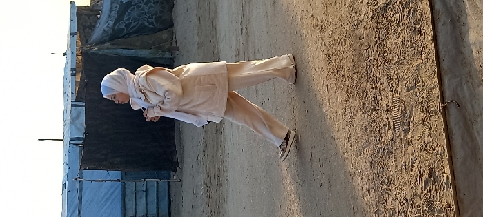 A young female individual is depicted walking from left to right across unpaved, sandy terrain in Khan Yunis, Palestinian Territory. She wears a light-colored hijab, a loose-fitting light beige jacket with two front pockets, and matching wide-leg trousers. Her left foot is visible, shod in a dark sandal. Her hands are clasped in front of her chest, and her head is turned to the right. The ground is dry, light brown, and marked with numerous footprints and tire tracks. In the background, several temporary structures are visible under a bright, clear sky. These include a large white tarp tent to the left and multiple darker, possibly canvas or tarp structures to the right, some with visible patterns, all supported by poles. Long shadows are cast across the ground, indicating a low sun angle.