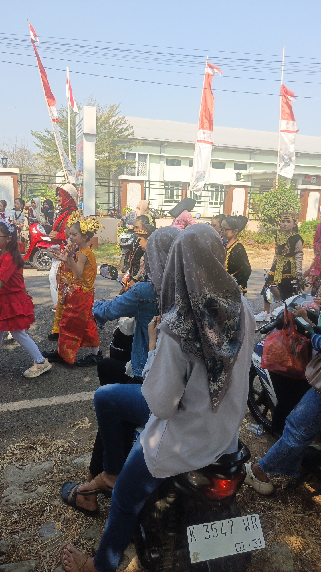 In a sunlit outdoor scene, a group of people, primarily women and children, are gathered along a paved road. Several are dressed in traditional attire, suggesting a parade or cultural event. A young girl in a vibrant yellow and red outfit with a gold headdress is the central focus, appearing to perform or pose. Other children are also in costume, some wearing headscarves and traditional jackets.
Several motorcycles are parked or moving, with individuals seated on them, some watching the proceedings. The background features a building with green trim and a fence, along with tall poles adorned with red and white flags, indicative of Indonesian national flags. The ground is a mix of asphalt and dry grass. The overall atmosphere is festive and communal, with a clear sense of a local celebration taking place during daylight hours.