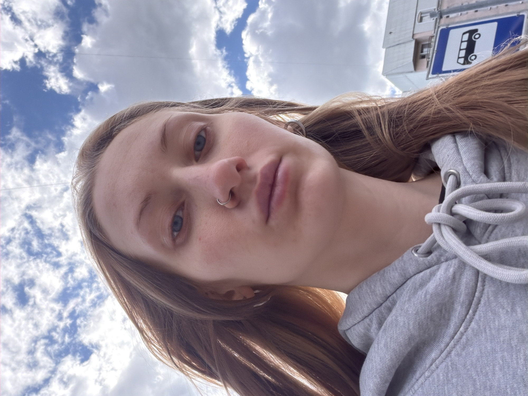 A low-angle, close-up selfie shows a young woman with long, light brown hair and blue eyes. She is wearing a grey hoodie and has a nose ring. The background is a cloudy sky with some buildings visible on the right, including a blue and white bus stop sign. The woman is positioned in the foreground, with her face slightly angled towards the camera. The overall scene appears to be outdoors in an urban environment. Based on the bus stop sign design, it is likely a public transportation stop in Saint Petersburg, Russia.