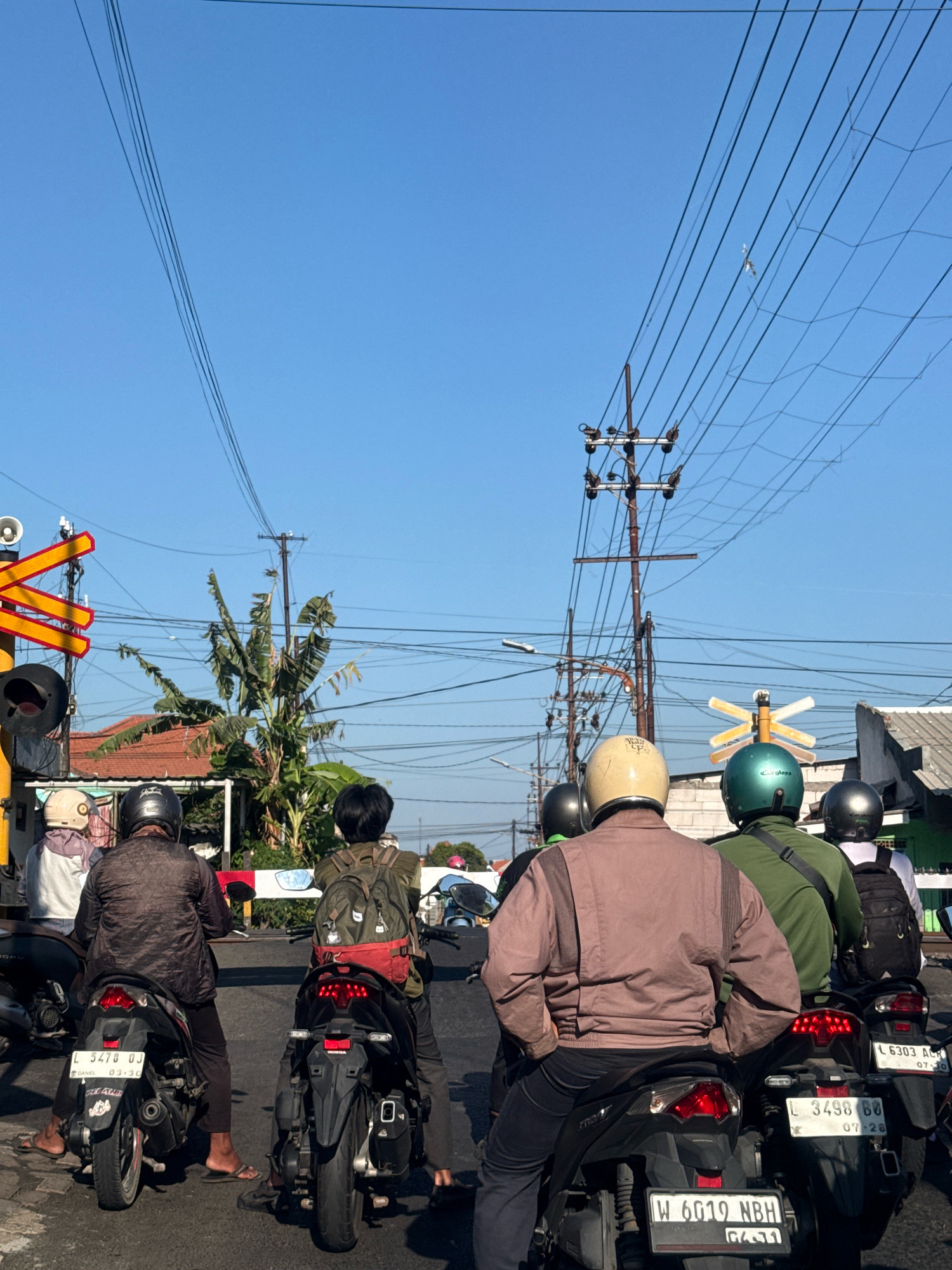 In Surabaya, Indonesia, several people on motorcycles are stopped at a railway crossing. The sky is clear and blue, with numerous power lines crisscrossing overhead. A traffic signal with red and yellow lights is visible on the left. People are wearing helmets, and some have backpacks. The motorcycles are parked on a paved road. Buildings and banana trees are visible in the background.
