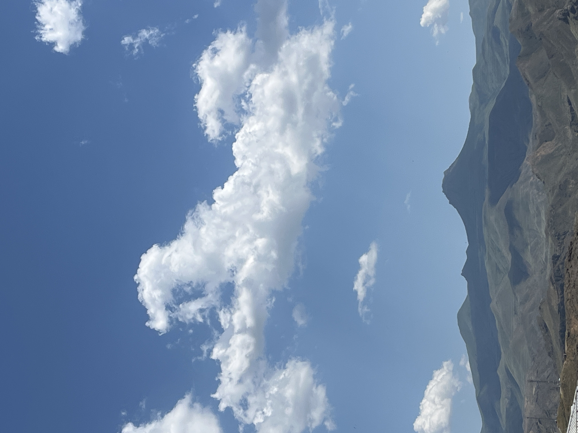 The image shows a landscape view with a focus on the sky and a mountain range. The sky is a clear blue with several fluffy white clouds scattered throughout. The mountains are a dominant feature, appearing in the right half of the frame. They are steep and rugged, with visible textures and shadows suggesting a sunny day. The lighting indicates it is daytime, likely late morning or early afternoon, given the angle of the shadows on the mountains. The weather appears to be fair and clear. The scene is natural and appears to be an outdoor setting, possibly a remote or mountainous region. There are no people or identifiable objects, other than the natural elements of clouds and mountains. There is no visible text in the image. The image is oriented vertically.