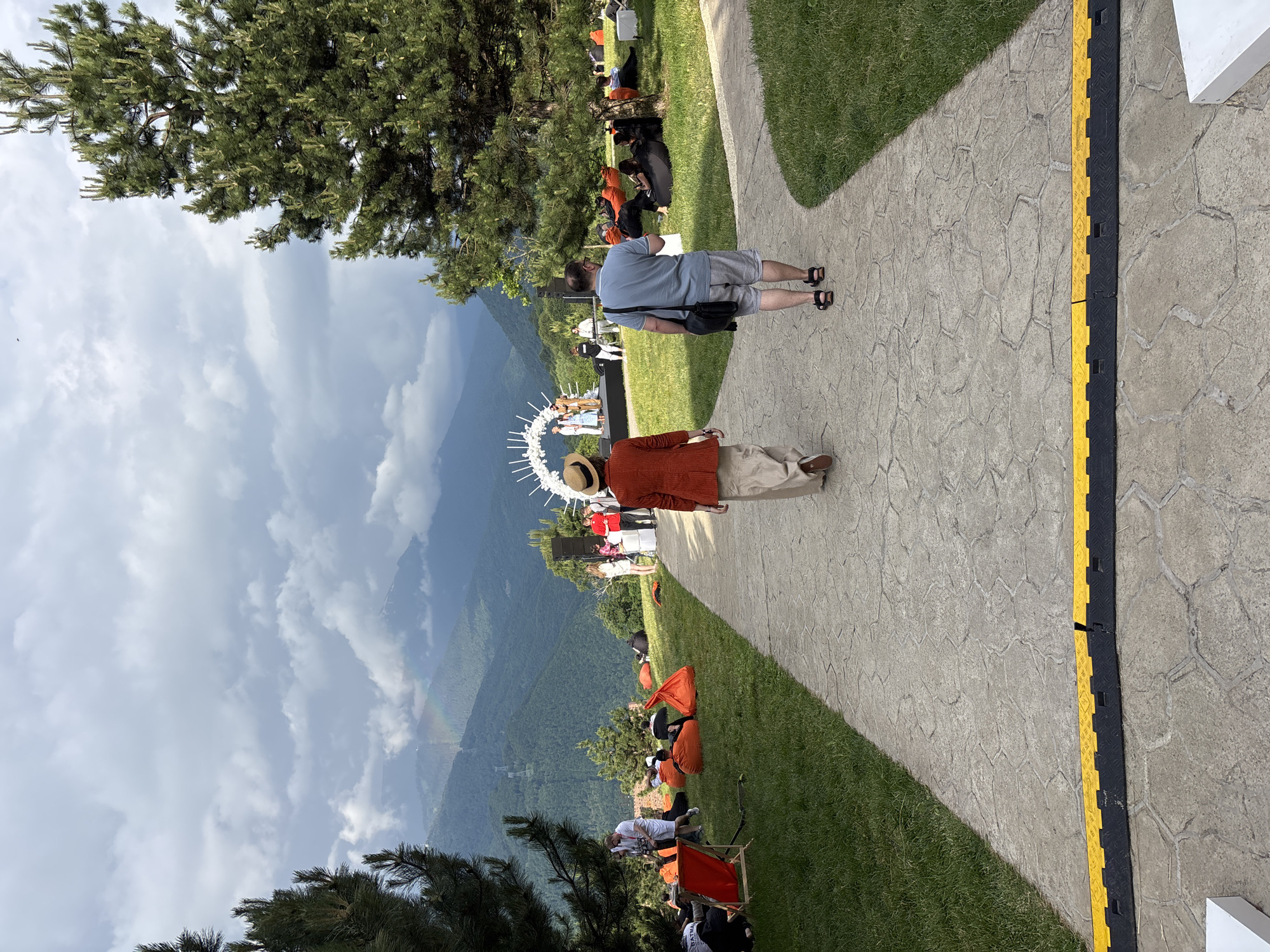This media file captures an outdoor event in Estosadok, Russia, set against a backdrop of lush green mountains under a cloudy sky with a visible rainbow. The scene appears to be a gathering or ceremony, possibly a wedding, with a decorative archway visible in the foreground.
Several people are present, with two individuals prominently featured in the foreground walking along a paved pathway. One person is wearing a blue t-shirt and grey shorts, carrying a dark bag, while the other is in an orange jacket, a straw hat, and khaki pants. Further back, a group of people are seated on the grass, some on orange beanbag-like chairs, facing a stage or platform where a ceremony is taking place. There are also individuals standing and observing the event.
The setting suggests a natural park or a venue with a scenic view. The time of day appears to be daytime, with natural light illuminating the scene. The weather is mixed, with clouds suggesting potential for rain, yet a rainbow indicates some sunlight. The terrain is sloped, with a well-maintained lawn and a paved walkway. A yellow and black cable protector runs along the edge of the walkway. The overall mood seems relaxed and celebratory. There is no visible text in the media file.