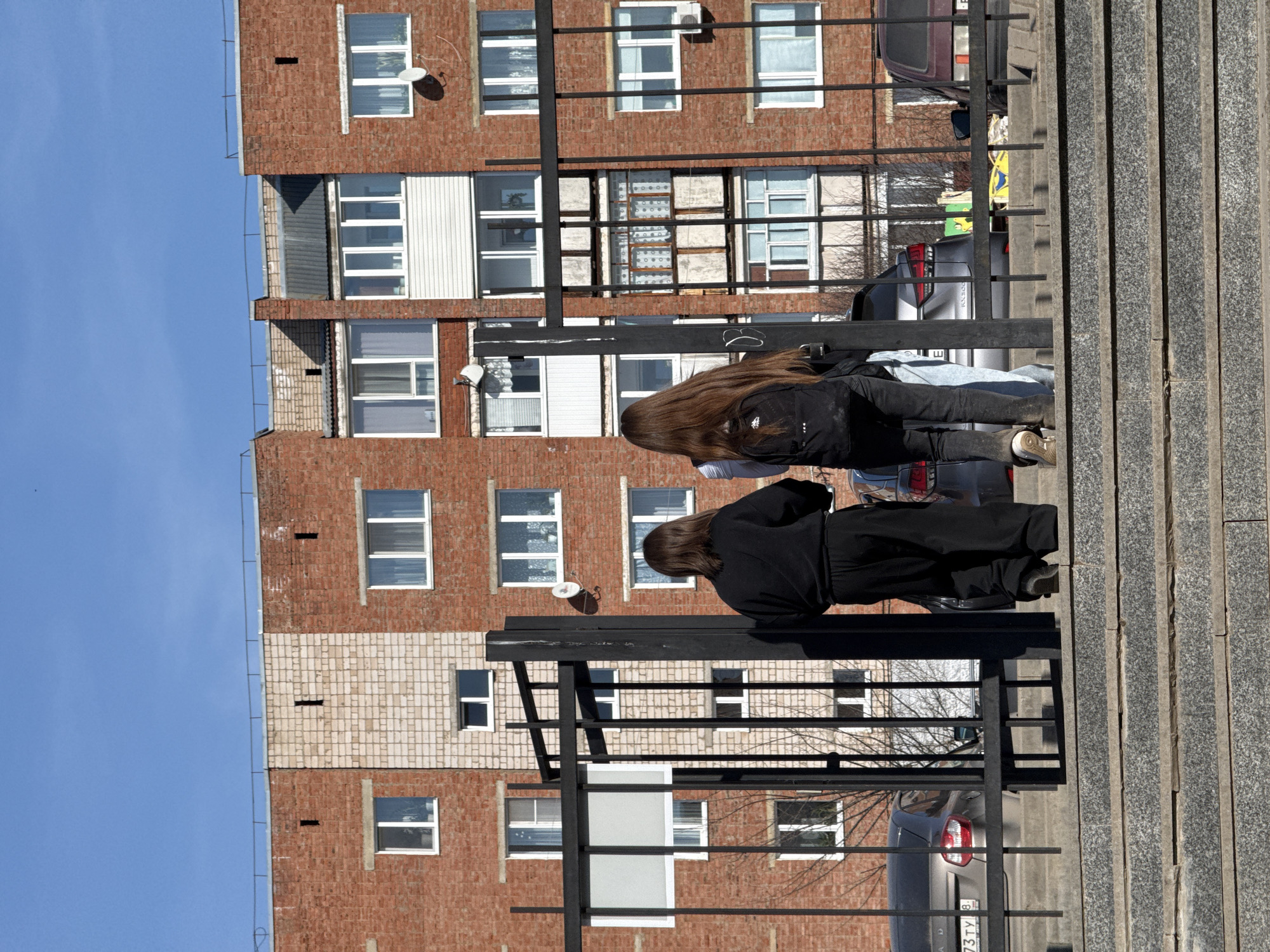 Two people are standing on a sidewalk next to a metal railing in what appears to be an urban setting in Votkinsk, Russia. The person in the foreground is wearing a black outfit. The second person is wearing lighter colored pants and a jacket. Behind them is a brick building with white-framed windows and satellite dishes. Cars are parked along the street. The sky is clear and blue.