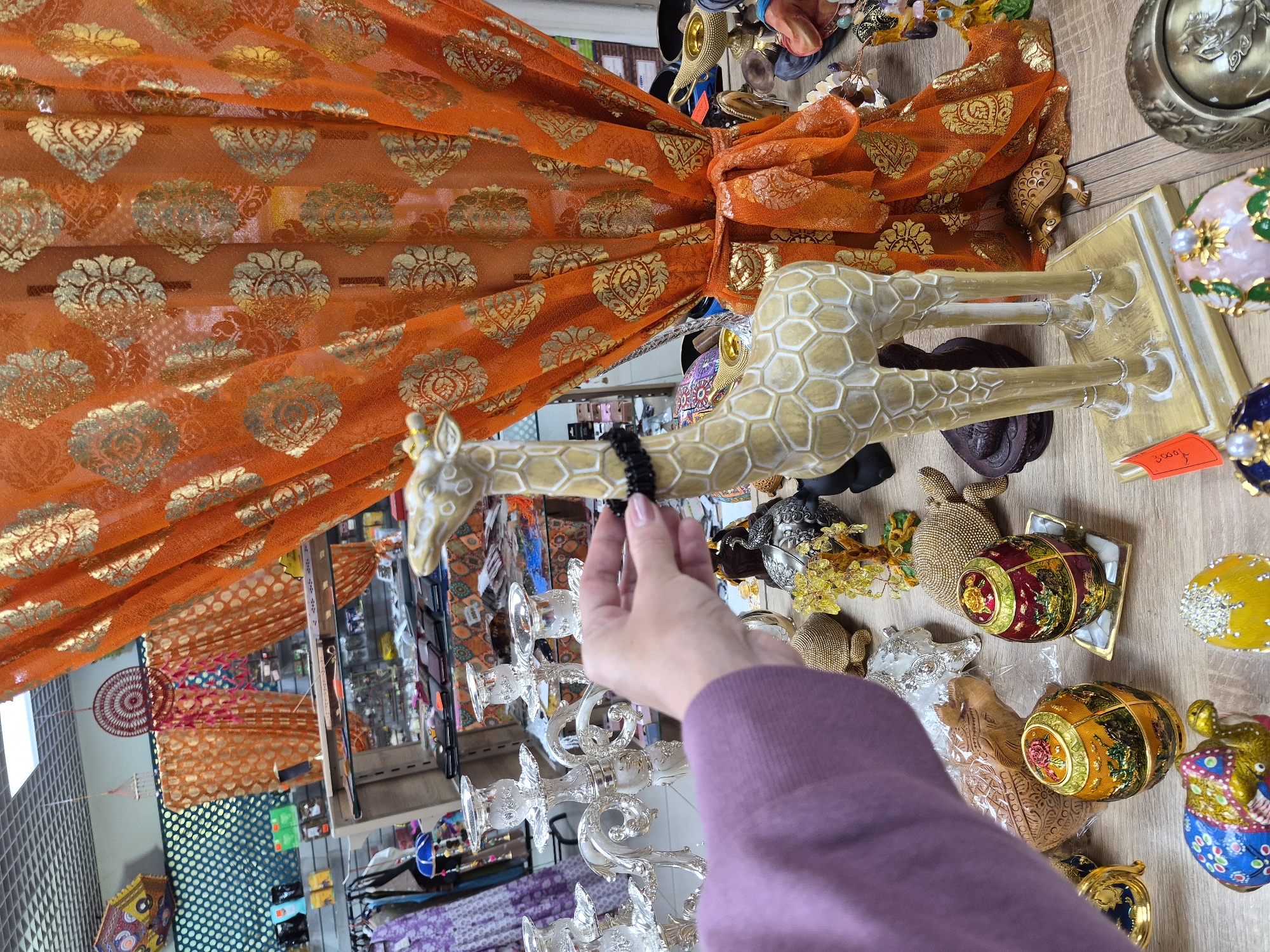 In Yekaterinburg, Russia, a person's hand, wearing a purple sweater, is adjusting a small black beaded necklace on the neck of a decorative giraffe figurine. The giraffe stands on a wooden surface amongst various decorative items, including ornate eggs, candle holders, and other animal figurines like turtles and elephants. An orange fabric with gold floral patterns is draped behind the giraffe, creating a backdrop with a store setting visible in the background.