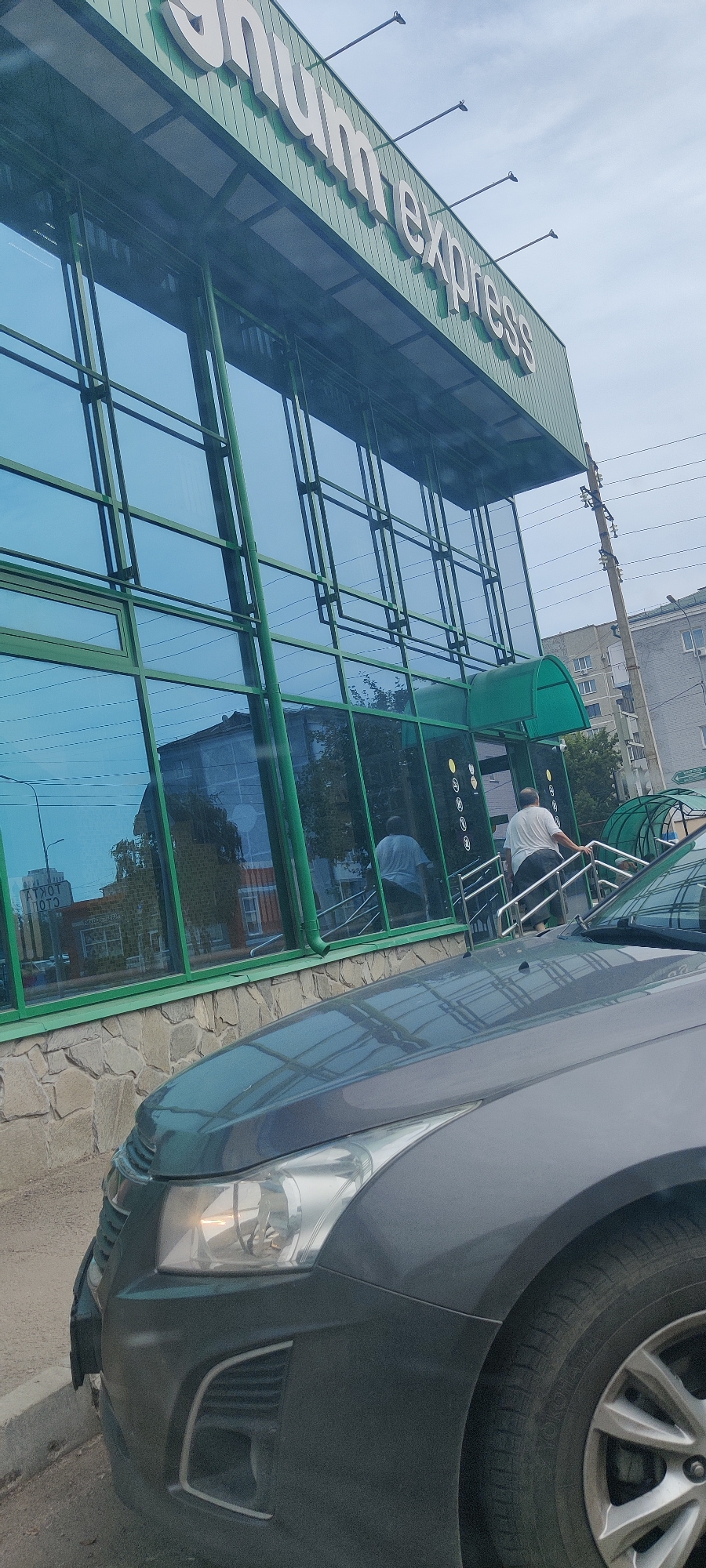 The image shows the facade of a business with large blue-tinted windows and green trim. A sign with white lettering reads "shum express" at the top of the building. A grey car is parked in front of the building, partially obscuring the lower portion of the facade. A man in a white t-shirt and dark pants is walking up a set of stairs to the right of the entrance. The street in front of the building appears to be paved. The sky is overcast. The building is likely located in a commercial or mixed-use area of the city of Petropavl, Kazakhstan.