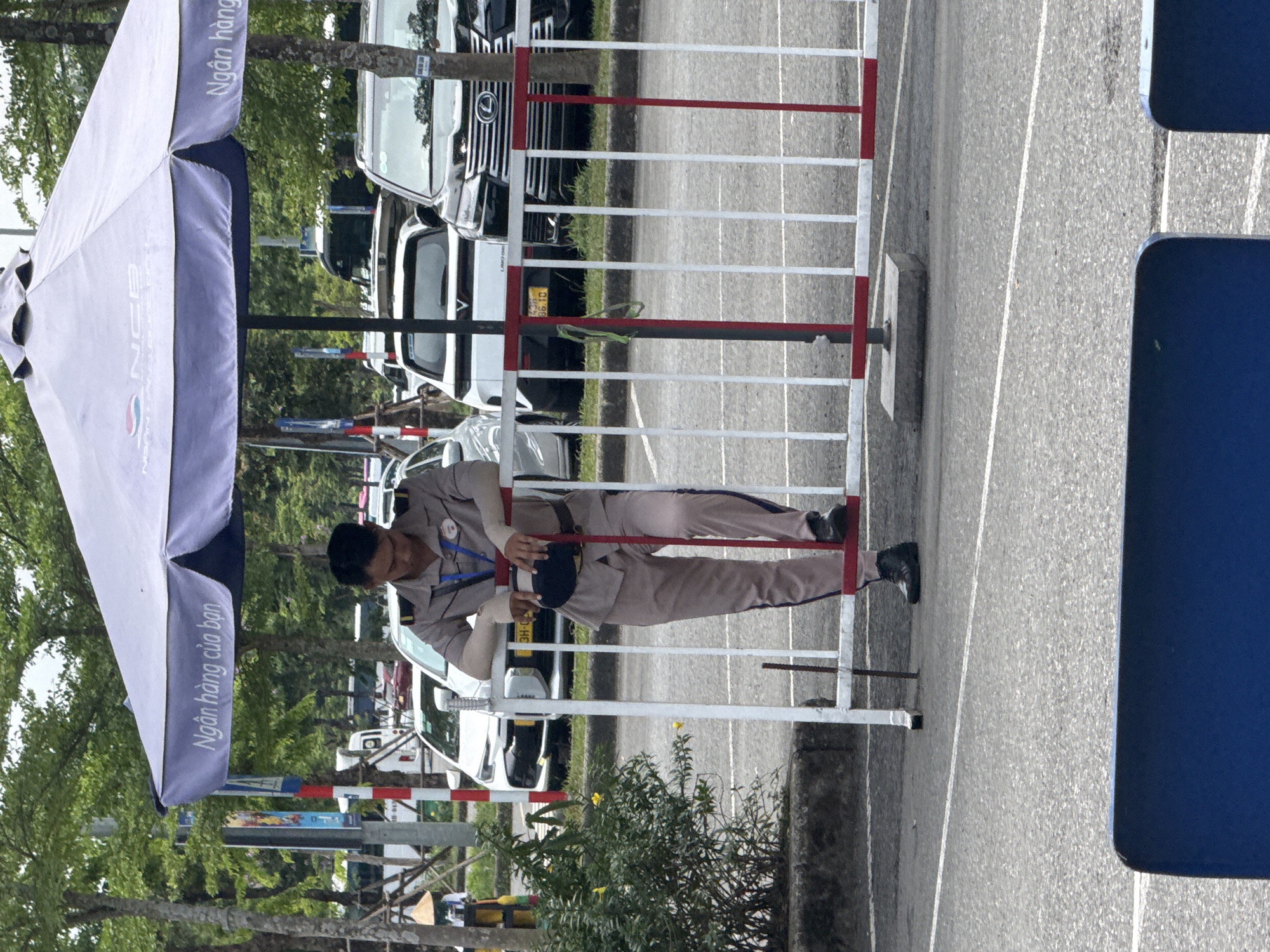 A man in a tan uniform is resting on a white and red metal barrier in an outdoor environment. Behind him, there is a row of parked white cars. The environment appears to be a parking lot with trees in the background. An umbrella stands on the left with Vietnamese script on its side. The barrier is situated on a concrete base on an asphalt road. The man appears to be looking down, possibly at something he is holding. Based on the signage and vehicle license plates visible in the image, the location is likely in the city of Đà Nẵng, Vietnam.