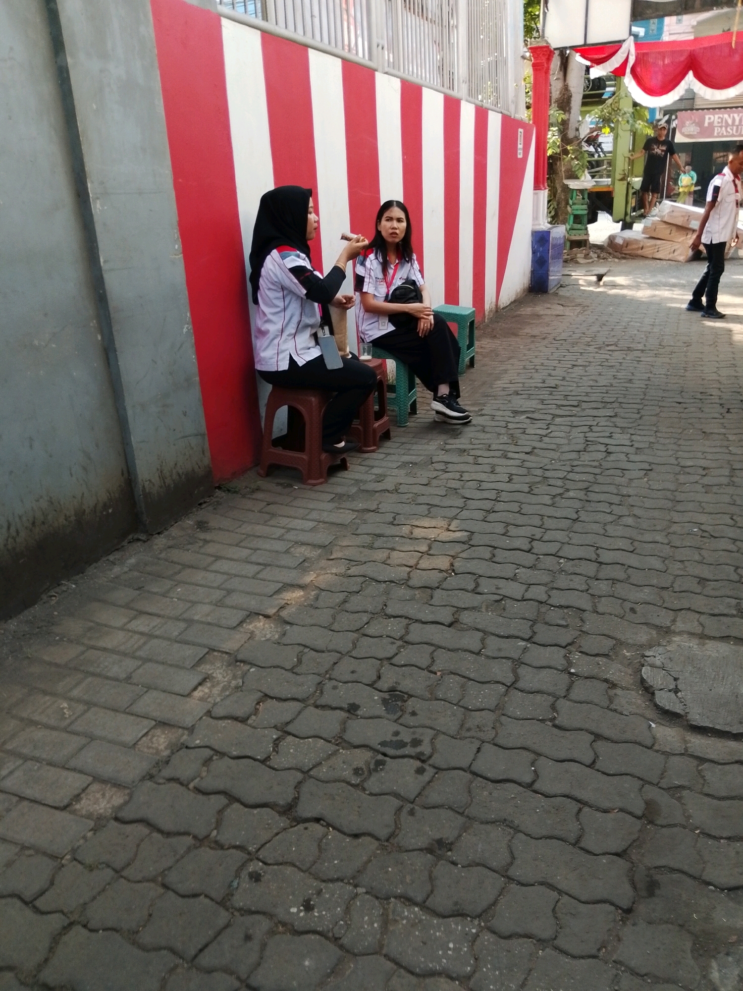 Two young women are sitting on plastic stools on a paved walkway. The woman on the left wears a black hijab and a white and red uniform. She is holding a dark object in her hand and appears to be speaking. The woman on the right also wears a white and red uniform and has her hands clasped in her lap. She is looking towards the woman on the left. Behind them is a wall painted with alternating red and white vertical stripes. In the background, other people are visible, including a man in a white shirt and black pants. The walkway is made of interlocking grey pavers. The scene appears to be an outdoor area in a city, likely in Pasuruan, Indonesia, given the provided context.