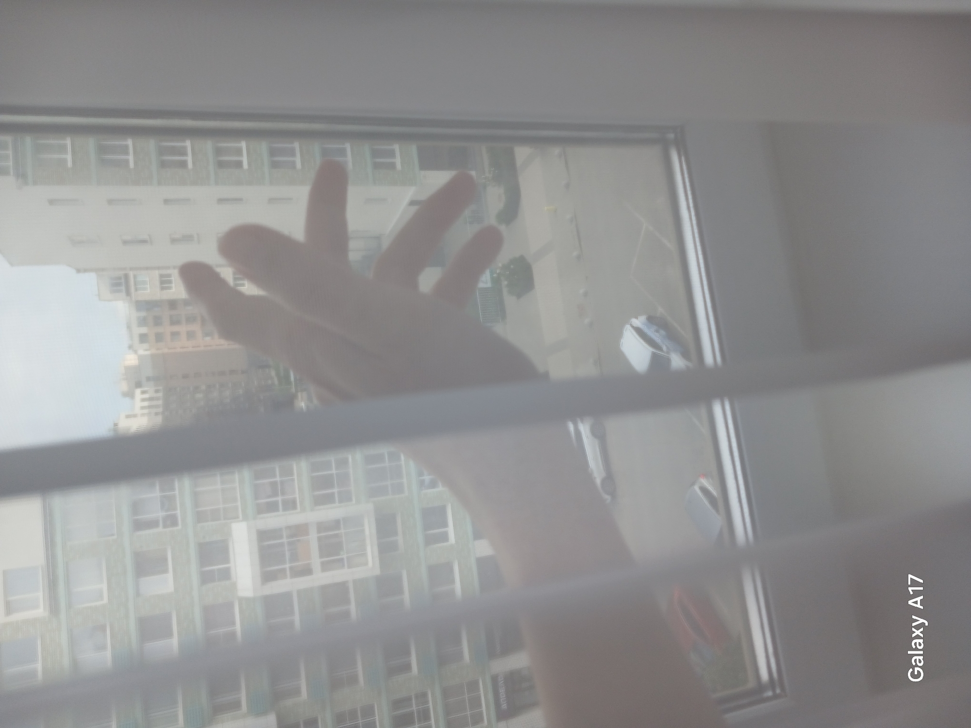 A view from a window through horizontal blinds shows a hand reaching out. The building across the street has a facade with many windows. A parking lot is visible with multiple cars. The phone's watermark indicates the photo was taken with a Galaxy A17. The scene appears to be Yekaterinburg, Russia.