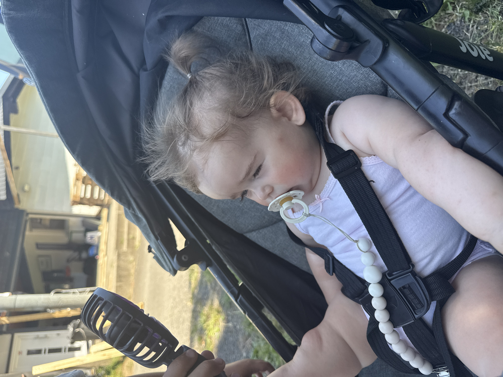 In Beltershain, Germany, a close-up shot captures a baby girl seated in a stroller. The baby, with her hair tied in pigtails, wears a white tank top and has a pacifier in her mouth. A string of white beads is attached to the stroller's harness, leading to the pacifier. To the left, a person's hand holds a black portable fan, directing its airflow towards the baby. The background reveals a residential area with a house and wooden structures. The scene is lit by natural daylight.