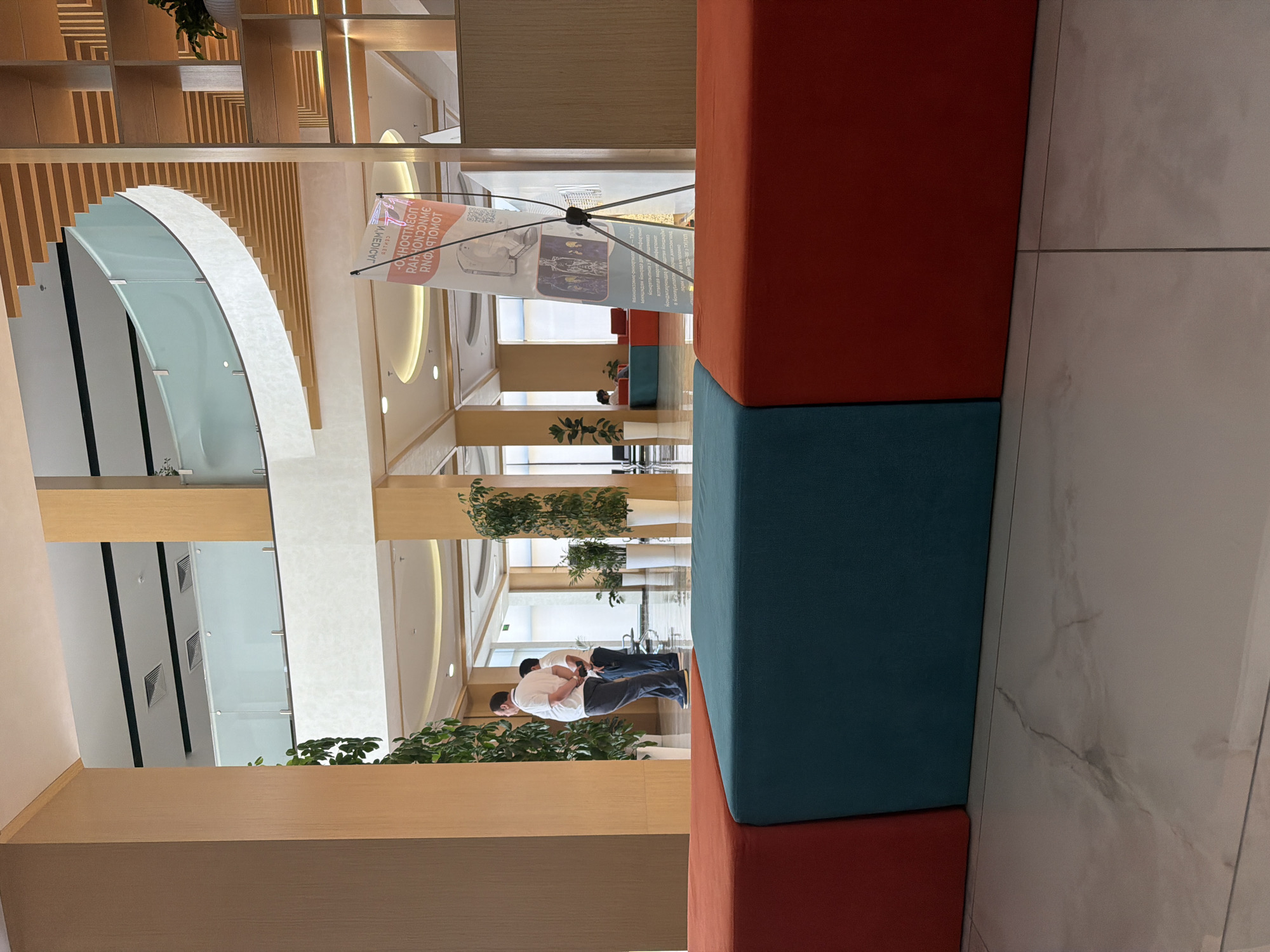 The image shows the interior of a modern building, likely a medical center or hospital, with a focus on a brightly colored, modular seating area in the foreground. Two men in casual attire are standing and looking towards the left side of the frame, near a large potted plant. The background reveals a multi-level interior with a curved balcony, wooden architectural elements, and natural light filtering in from windows. A promotional banner, which appears to be for a medical facility with text in a foreign language (likely Uzbek or Russian, given the presumed location of Tashkent, Uzbekistan), is displayed on a tripod. The flooring is polished marble.