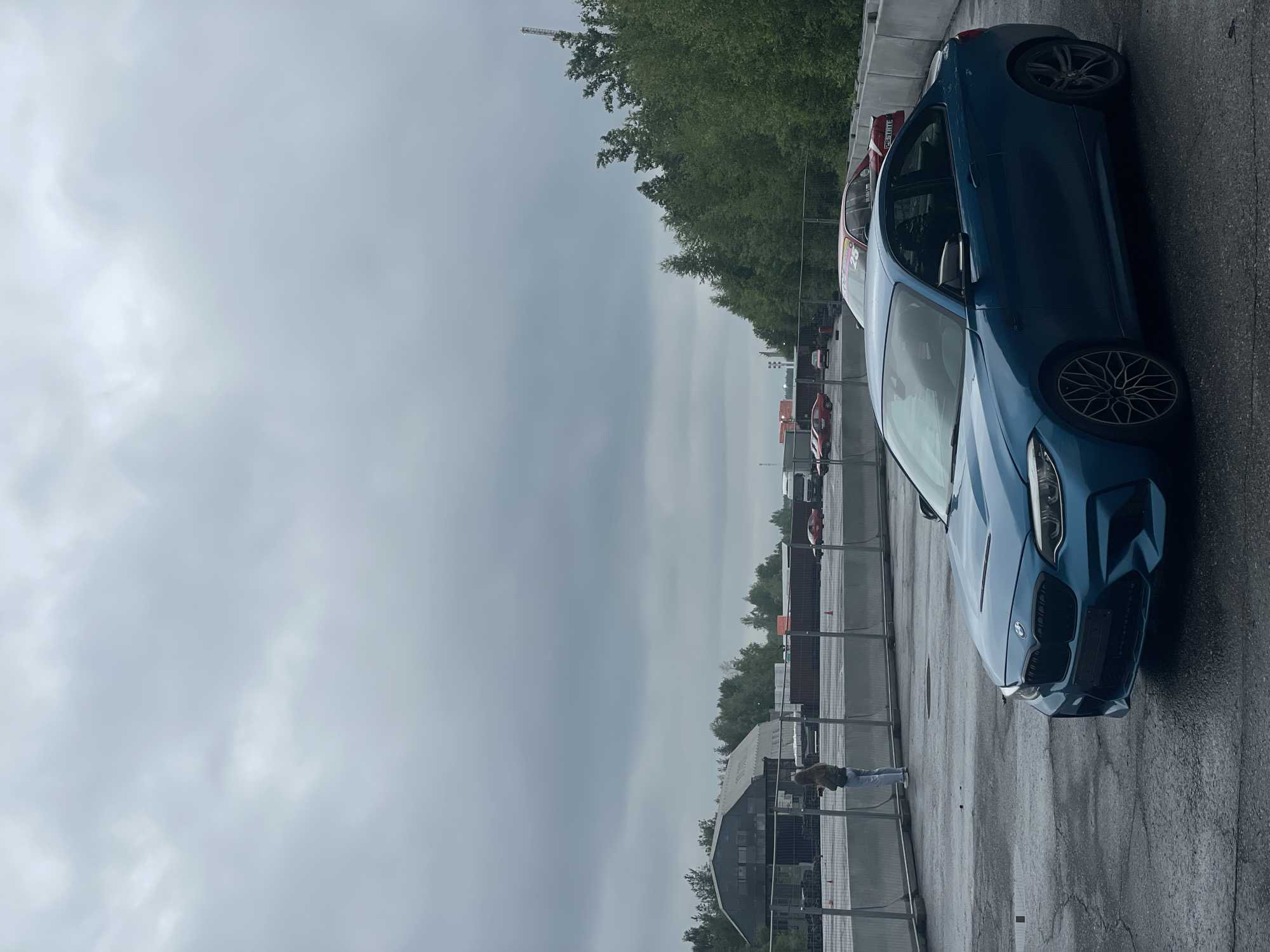 In the city of Kovalyovo, Russia, an overcast sky looms over an outdoor setting, possibly a race track or industrial area. In the foreground, a teal BMW M2 is parked on a wet asphalt surface. The car is positioned at an angle, with its front and driver's side visible. To the left of the BMW, a person stands near a metal fence, facing away from the camera. Behind the person and the fence, a large, grey industrial building with a curved roof is visible. Several shipping containers are stacked behind the fence, and more vehicles, including red cars and a white truck, are present in the background. Dense green trees line the right side of the scene.