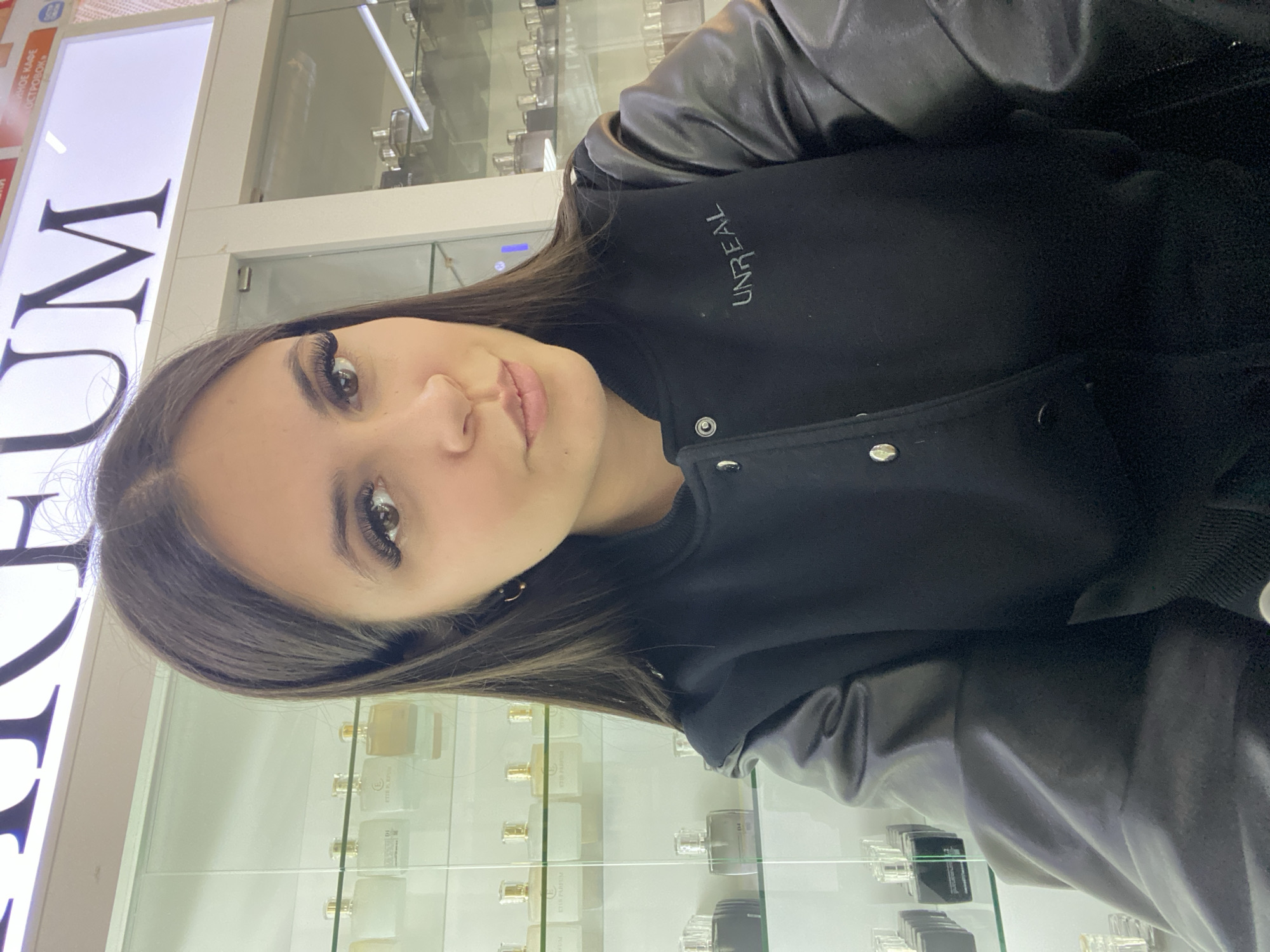 A young woman is captured in an indoor setting, likely a retail store in Chaikovsky, Russia. She has long, dark hair and dark eyes with prominent eyelashes, and she is looking directly at the camera with a neutral expression. She wears a black jacket with contrasting leather-look sleeves, and the word "UNREAL" is visibly embroidered on the chest.
The background features brightly lit display cases filled with numerous perfume bottles, indicating a fragrance or cosmetics store. A large, illuminated sign is partially visible behind her, displaying the letters "UM" (likely part of "ПАРФЮМ," meaning Parfum in Russian, which is also seen on some perfume bottles as "ETD PARFUM"). Other visible Cyrillic text on the sign includes "НОЕ КАФЕ" (coffee shop) and "ОСТРОВОМ" (island). The artificial lighting suggests the photo was taken indoors during any time of day. The image is rotated 90 degrees clockwise.