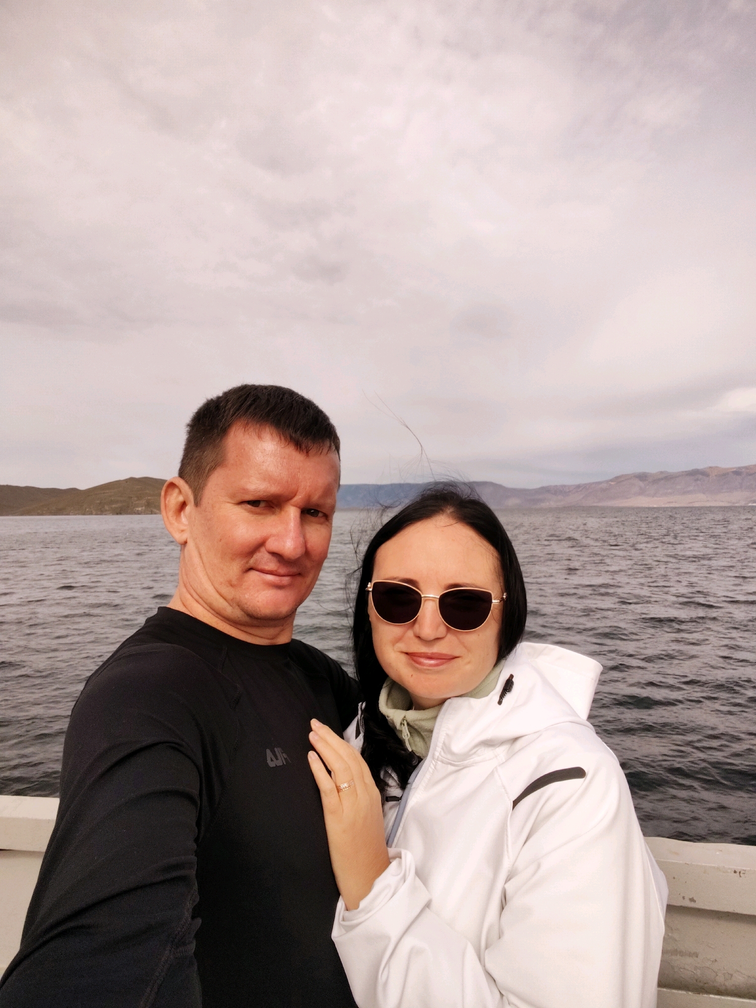 A medium shot captures a couple posing for a selfie on a boat with a body of water and hills in the background. The man, on the left, is facing forward with a slight smile, wearing a black long-sleeved shirt. The woman, on the right, is also facing forward, wearing sunglasses and a white hooded jacket. Her left hand rests on the man's arm, displaying a ring on her ring finger. The water is choppy and the sky is overcast with pale clouds. The location is identified as the village of Sakhiurta, Russia.