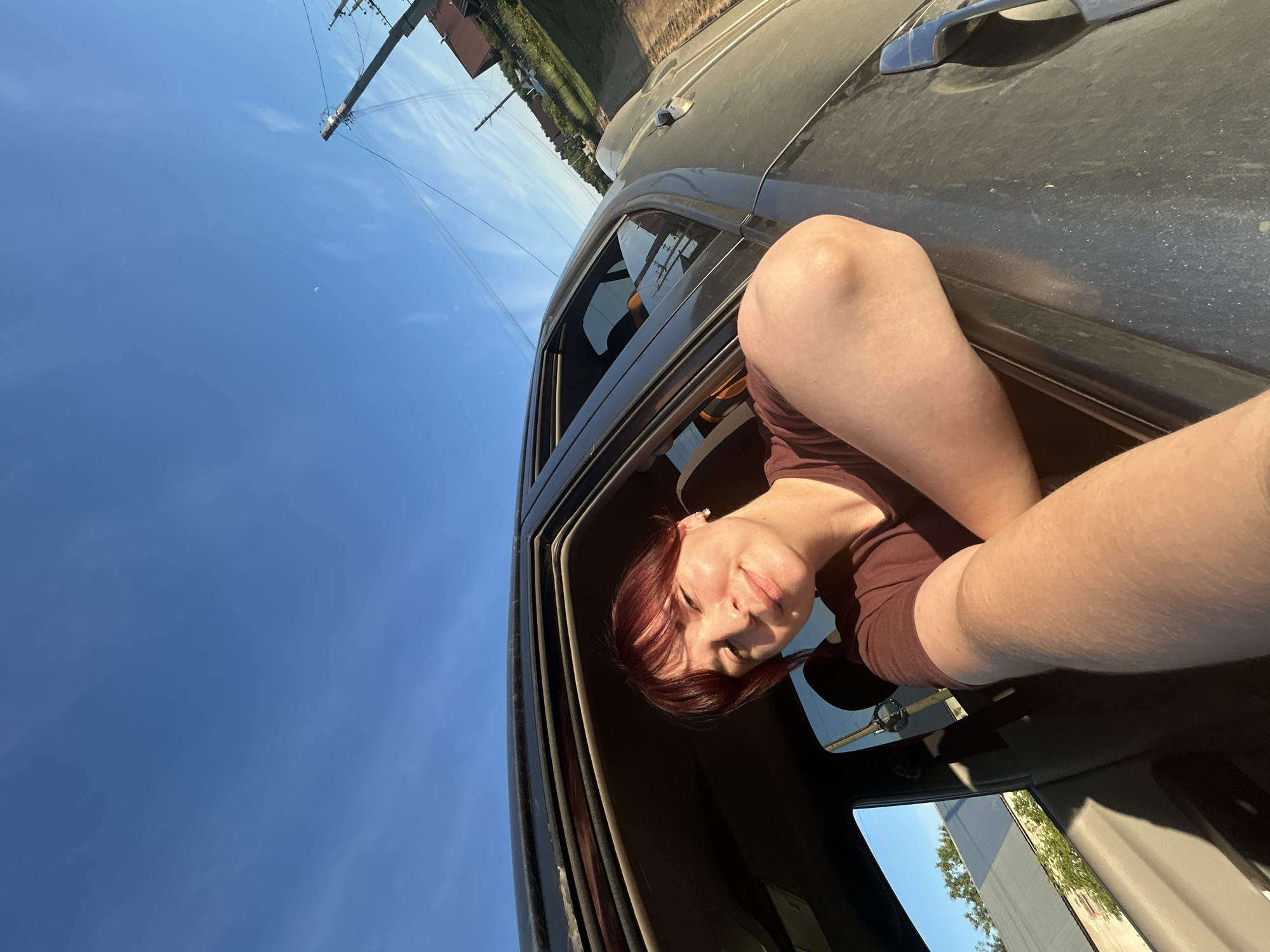 A young woman with reddish-brown hair and bangs is in the driver's seat of a dark-colored car, with her arm resting on the open window frame. She is smiling and looking towards the camera, her face illuminated by the setting sun. The background shows a clear blue sky with some wispy clouds, a utility pole with wires, and a hint of houses and trees, suggesting a rural or suburban setting. The image appears to be taken during golden hour. The location is identified as Fokino, Russia.