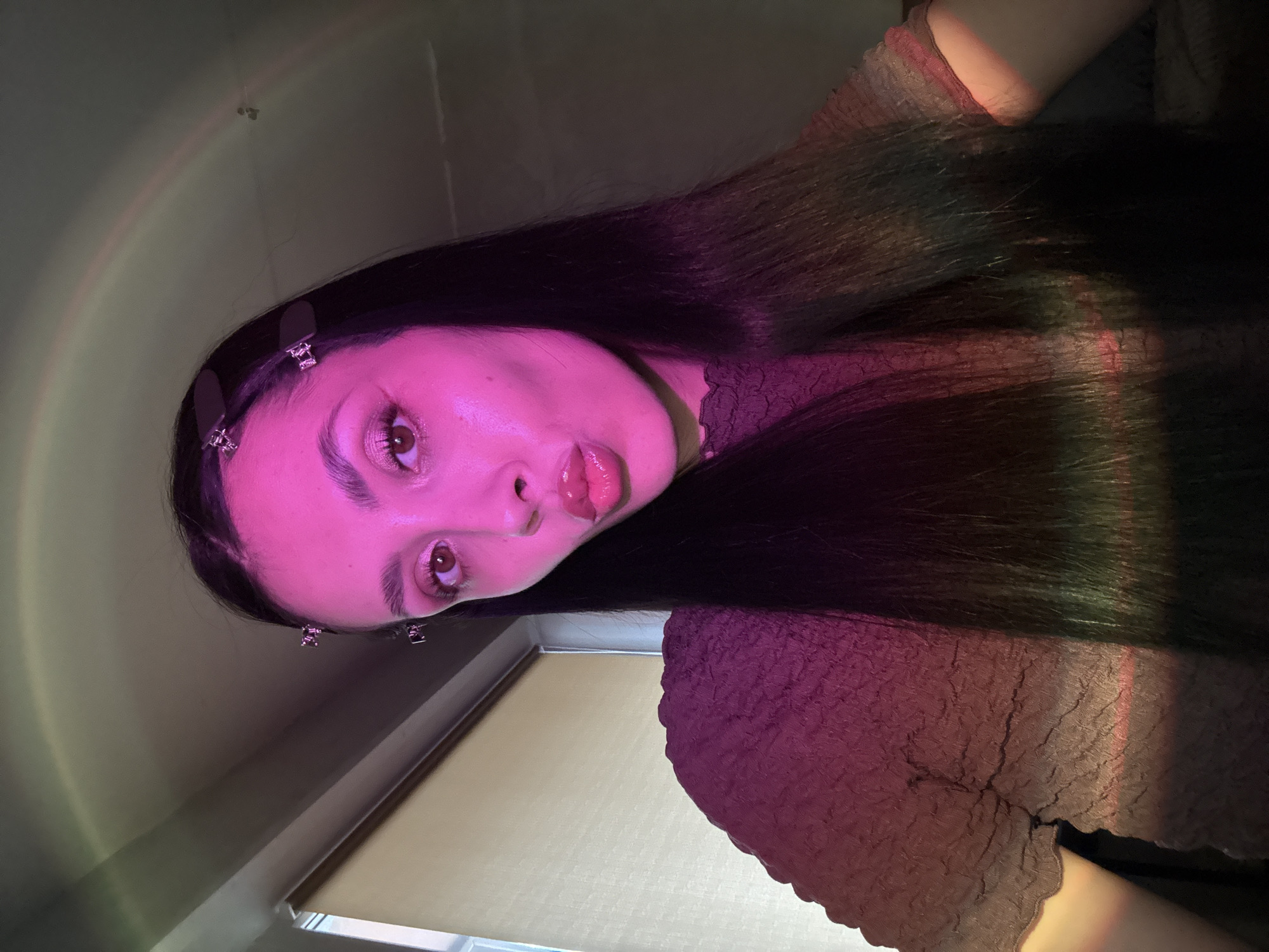 A woman with long, dark hair poses in a dimly lit interior. She is facing the camera and has light skin, prominent eyebrows, and glossy lips. Two small, decorative clips adorn her hair on the left side. Her clothing, a textured top, appears to be a dusky pink or mauve color. The background is nondescript, with a wall and what appears to be a closed window blind. A rainbow-like light effect is cast across the scene, illuminating the wall and part of the subject. There is no indication of the location being Votkinsk, Russia, nor any other details specific to location.