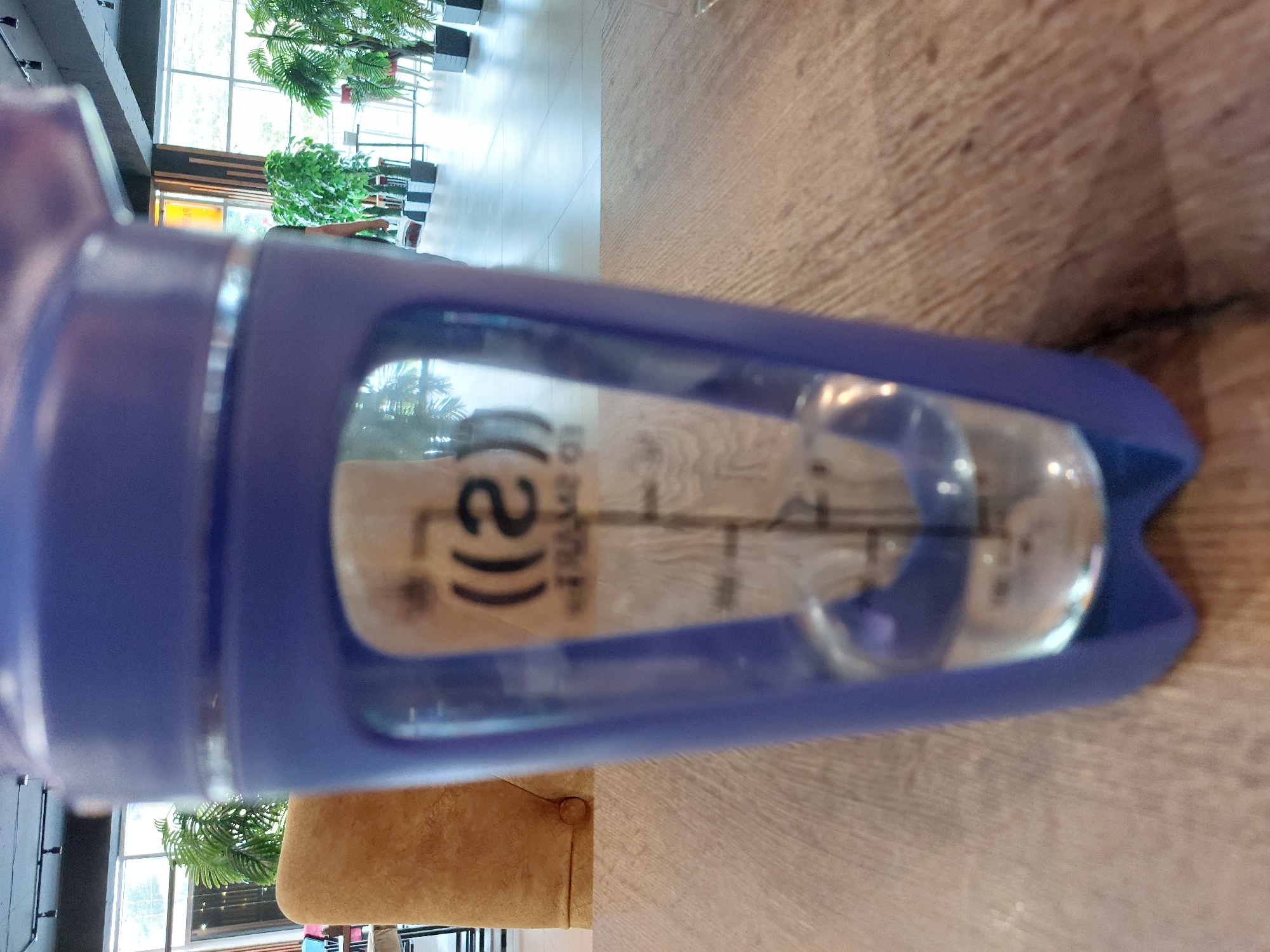 A close-up, eye-level shot shows a blurred, purple shaker bottle filled with a clear liquid, sitting on a wooden table. The bottle has a clear section with markings and what appears to be a logo with the text "((2)) TO SHAKE". In the background, a tan upholstered chair is visible on the left. Beyond the chair, through a large window, a bright, well-lit interior space is visible with plants and people. The flooring appears to be light-colored tile, and industrial-style lighting fixtures are attached to a dark ceiling. The overall setting suggests a modern, public indoor space, possibly a cafe or lounge. The image is not taken in Podkumok, Russia.