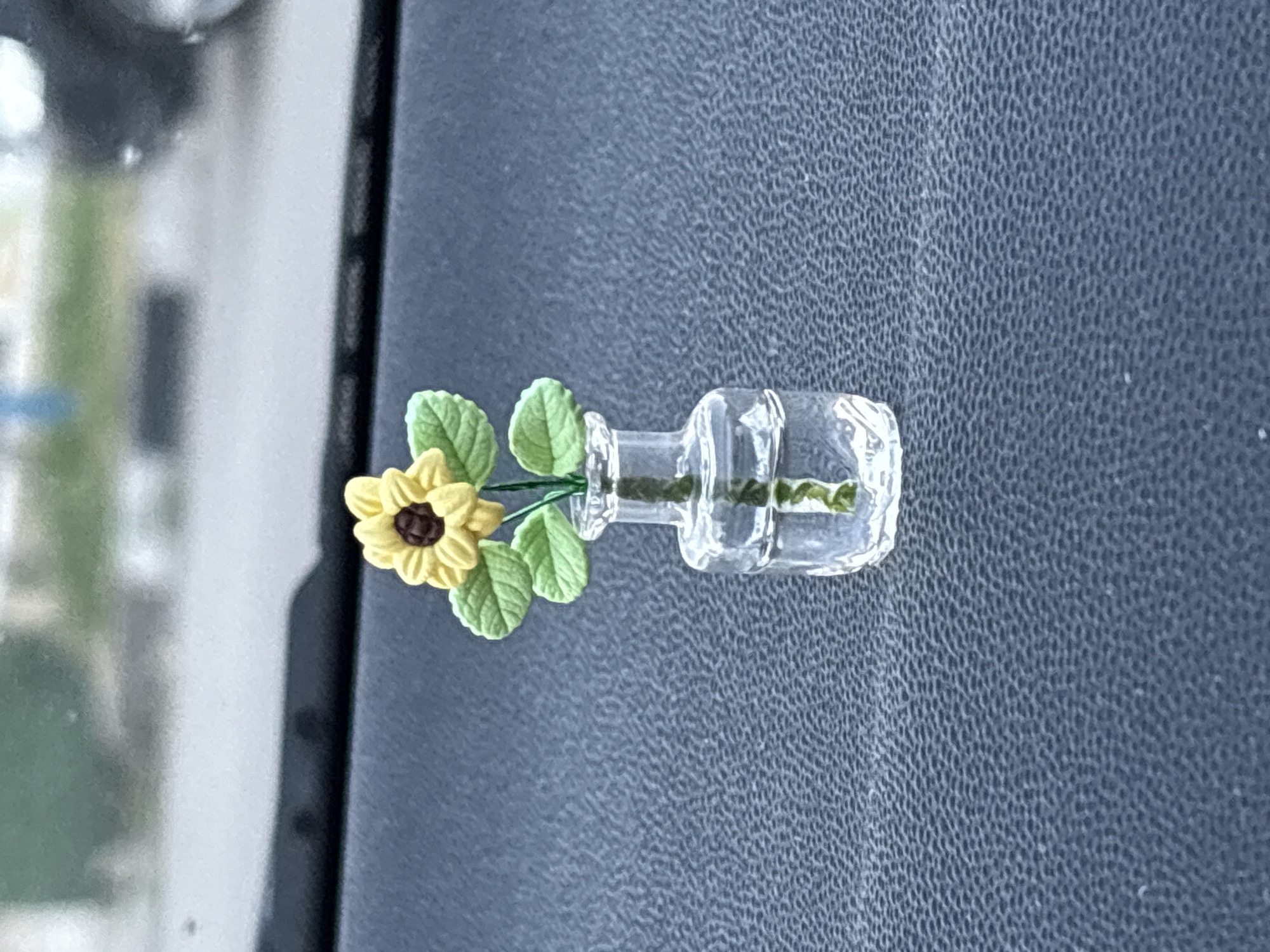 A miniature sunflower in a clear glass vase sits on a textured, dark surface, likely the dashboard of a vehicle, given the blurred background which suggests outdoor scenery and the interior of a car. The sunflower has yellow petals and a brown center, with green leaves. The vase contains a clear liquid and a green stem. The scene is lit from above. The location is identified as Tervel, Bulgaria, though no specific landmarks are visible to confirm this.