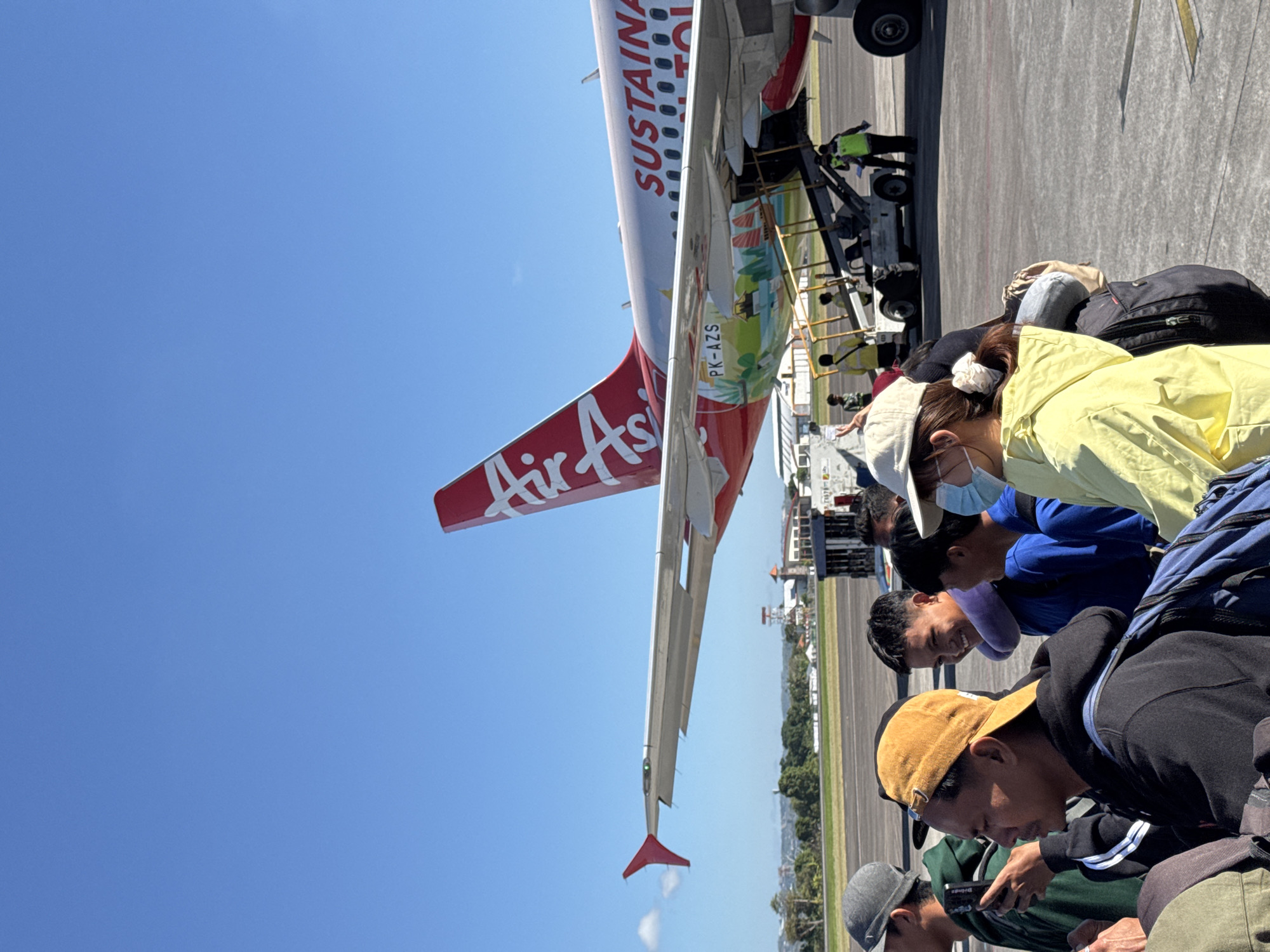 This media file captures an outdoor scene at an airport in Kuta, Indonesia. The primary subject is an AirAsia airplane, predominantly red and white, with "SUSTAINABLE" and "AirAsia" emblazoned on its fuselage. Its registration number, "PK-AZS", is also visible. The image also features a group of people, appearing to be passengers, gathered on the tarmac. They are dressed in casual attire, with some wearing hats and backpacks. The activity depicted is likely boarding or disembarking an aircraft, given the proximity to the plane and the presence of what appears to be a mobile staircase or jet bridge.
The scene is set under a clear, bright blue sky, suggesting it is daytime with good weather. The sun is high, casting strong shadows, indicating it is likely midday. The ground is paved with asphalt, characteristic of an airport runway or apron, with faint yellow lines visible. In the background, there are glimpses of airport buildings and lush green trees, typical of a tropical location like Indonesia.
Several individuals are looking at their phones, suggesting a period of waiting. One person in the foreground wears a yellow jacket and a face mask, and another person is wearing a light blue face mask. The emotions conveyed are neutral, with individuals appearing to be calmly waiting or engaged with their devices. The overall impression is one of a typical travel day, with passengers preparing for their flight.