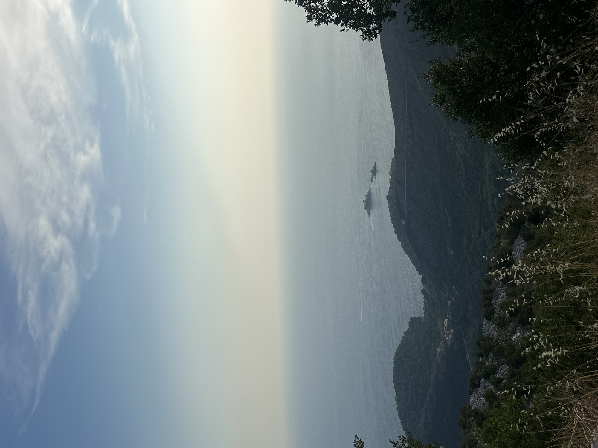 This is a high-angle, full shot of a coastal landscape from a mountainous vantage point, overlooking a body of water that appears to be the Adriatic Sea. The sky is a clear, pale blue with scattered wispy clouds, transitioning to a hazy yellow near the horizon. Below the sky, the sea is a muted blue-grey, with two small, rocky islands visible in the middle distance. The coastline features rolling, tree-covered hills that recede into the distance. In the foreground, there is dry, tall grass and shrubs in shadow, with some rocky outcrops. The overall impression is one of a serene, natural environment, likely in a Mediterranean climate, possibly near Novoselje, Montenegro, given its coastal and mountainous geography. There are no people or animals visible in the frame. The scene appears to be captured during daylight, likely late afternoon or early morning due to the soft light and haze.