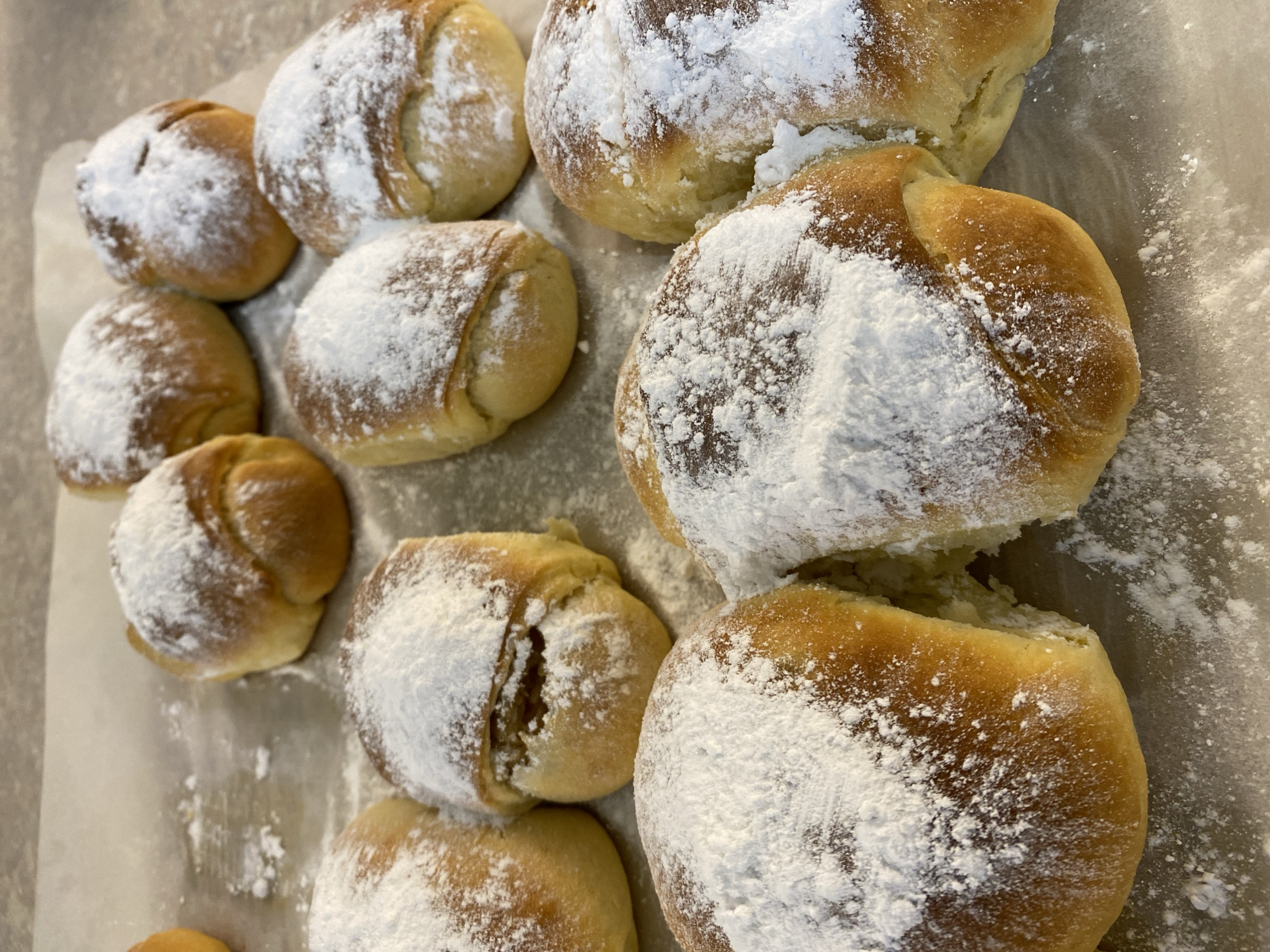 A top-down, close-up shot displays a batch of baked rolls, generously dusted with powdered sugar, arranged on a light-colored surface, likely parchment paper. The rolls vary slightly in size and shape, with some appearing rounder and smaller while others are larger and more irregular. The powdered sugar covers the tops of the rolls, creating a soft white contrast against the golden-brown crust of the baked goods. The image suggests a domestic setting, perhaps a home kitchen or bakery. There is no indication of the location in the city of Budva, Montenegro.