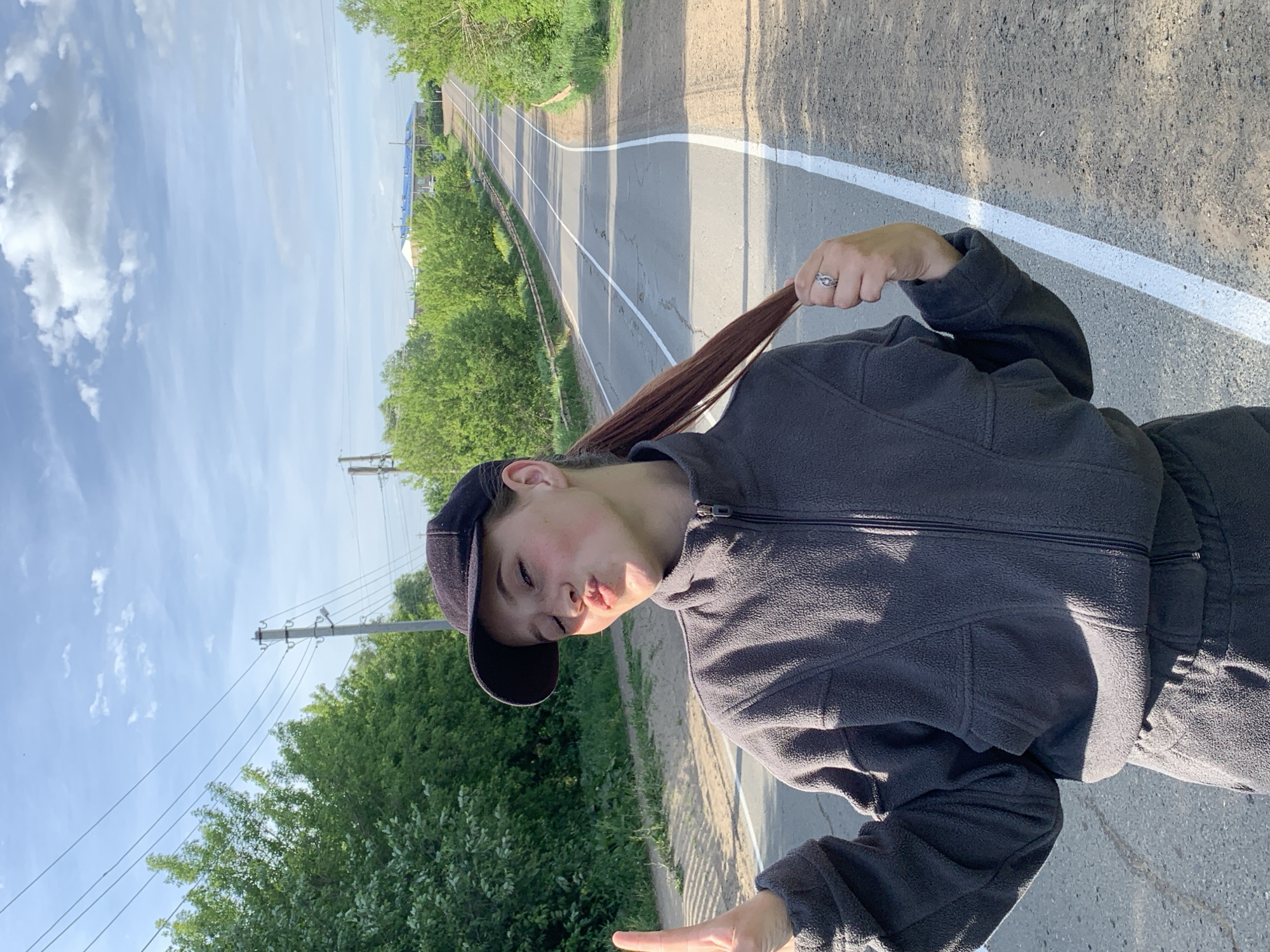 A young woman wearing a dark baseball cap and a fleece jacket is standing on a paved road with a white line running down the middle. She is holding a strand of her long brown hair up in her hand. The road stretches into the distance, flanked by green trees and vegetation on either side. Power lines are visible on the left side of the road. The sky is partly cloudy with blue patches. The location is in the city of Фоки, Russia.