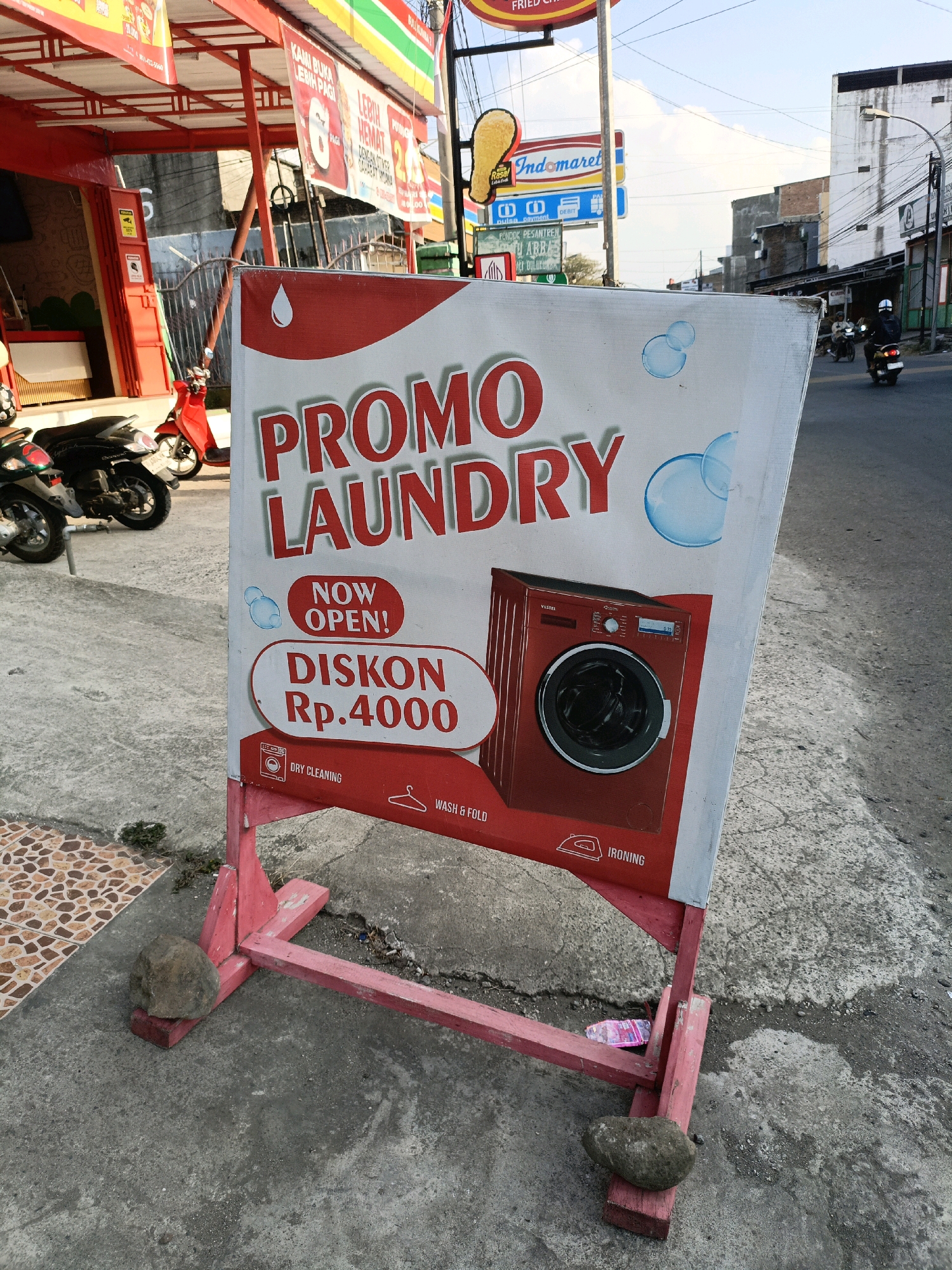 A promotional sign for a laundry service is displayed on a sidewalk in Bulukumba, Indonesia. The sign, made of white material with red lettering and graphics, advertises "PROMO LAUNDRY," "NOW OPEN!", and "DISKON Rp.4000". It features an image of a red washing machine and icons representing "DRY CLEANING," "WASH & FOLD," and "IRONING." The sign is supported by a pink wooden A-frame structure, with rocks used as weights to stabilize it on the concrete ground. In the background, there are motorcycles parked on the left, and on the right, a street scene with buildings, streetlights, and a motorcyclist riding past. The overall environment suggests an urban setting.