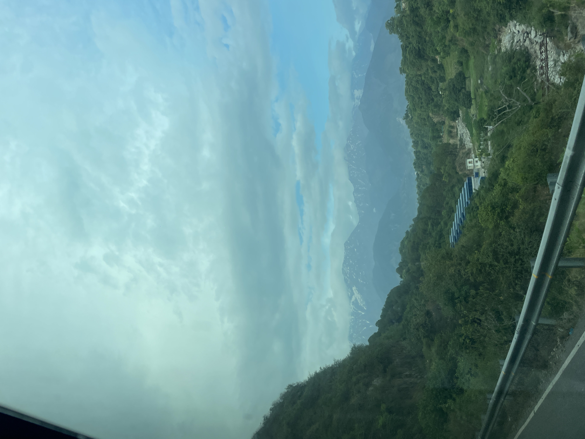 The image depicts a view from a vehicle, likely a car, looking out over a mountainous landscape near Upper Skoh, India. The sky is overcast with heavy clouds. In the distance, snow-capped mountains are partially obscured by mist. Lush green vegetation covers the foreground hillsides. A structure with a blue roof and other small buildings are visible amidst the foliage. A gray metal guardrail is present on the right side of the frame, indicating the edge of a road. A segment of the vehicle's interior is visible in the lower left corner.