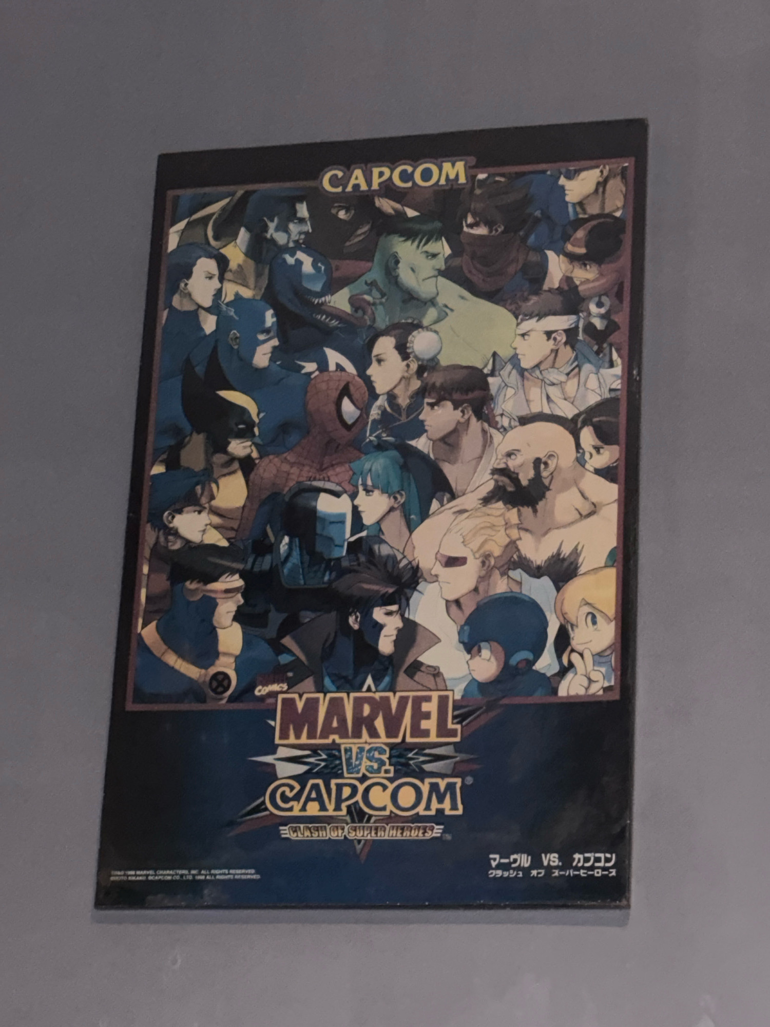 This image shows a poster for the video game "Marvel vs. Capcom: Clash of Super Heroes". The poster features a large illustration of many characters from both Marvel Comics and Capcom's fighting games, such as Wolverine, Spider-Man, Captain America, Ryu, Chun-Li, and Mega Man. The poster is mounted on a wall, and the scene appears to be indoors. The poster is the primary subject, and it is displayed in what looks like a home or personal space, not a commercial establishment. The background is a plain, gray wall. There is no discernible location within the city of Bekasi, Indonesia, or any other specific city. The image is a still photograph of the poster.