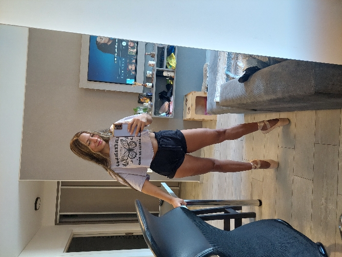A young woman, reflected in a mirror, strikes a ballet pose en pointe. She wears a t-shirt and shorts. The background includes a TV with shelving above, a gray sofa, a pet house, and a chair in the foreground. The floor is tiled. The woman holds a phone, presumably taking the photo in the mirror.