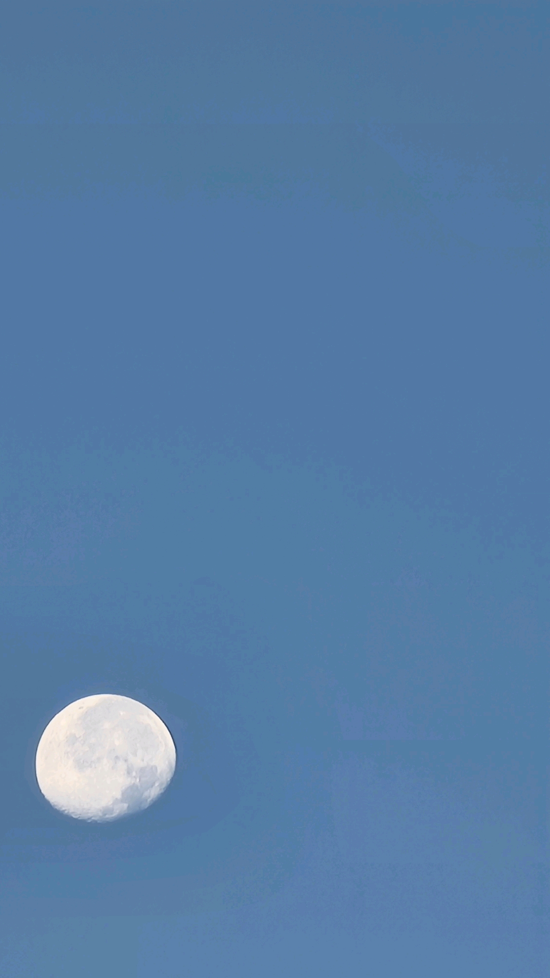 The image is a vertical, full-frame shot of a clear blue sky with a large, bright moon positioned in the lower left quadrant. The moon is illuminated and shows surface detail. The sky is a uniform light blue, with no clouds or other celestial bodies visible. The composition is minimalist, focusing solely on the moon against the vast expanse of the sky. There is no indication of the city of Sant Joan Les Fonts, Spain, in the image.