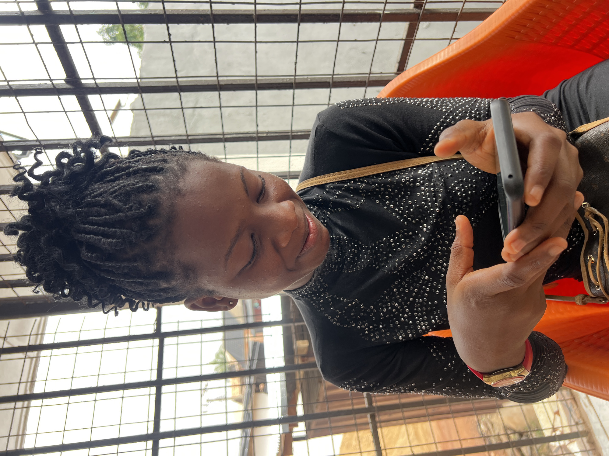 A young Black female is pictured indoors, seated on an orange plastic chair. She is looking down at her phone, which she holds in both hands. She has dark skin, black braided hair pulled up, and is wearing a black long-sleeved shirt adorned with numerous small, reflective embellishments. A red and gold watch is visible on her left wrist. A brown strap from a bag is draped across her chest. The background features a metal grate and a blurry outdoor scene with buildings and greenery, suggesting a location in Nkpor, Nigeria. The lighting is natural, coming from an unseen source to the left.