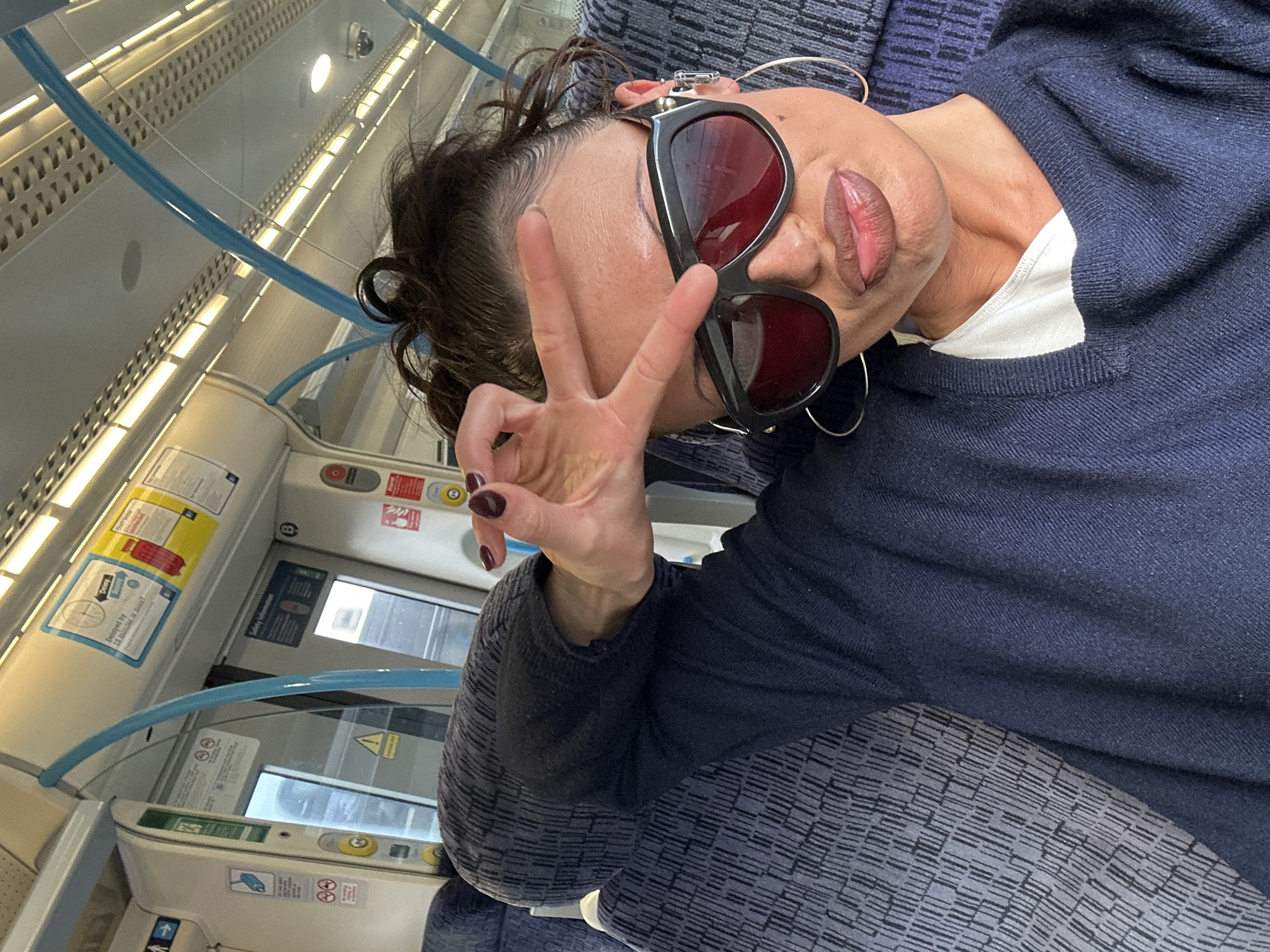 A woman wearing sunglasses and a navy blue sweater is on a train. She is making a peace sign with her left hand. The train is in the city of London, United Kingdom.