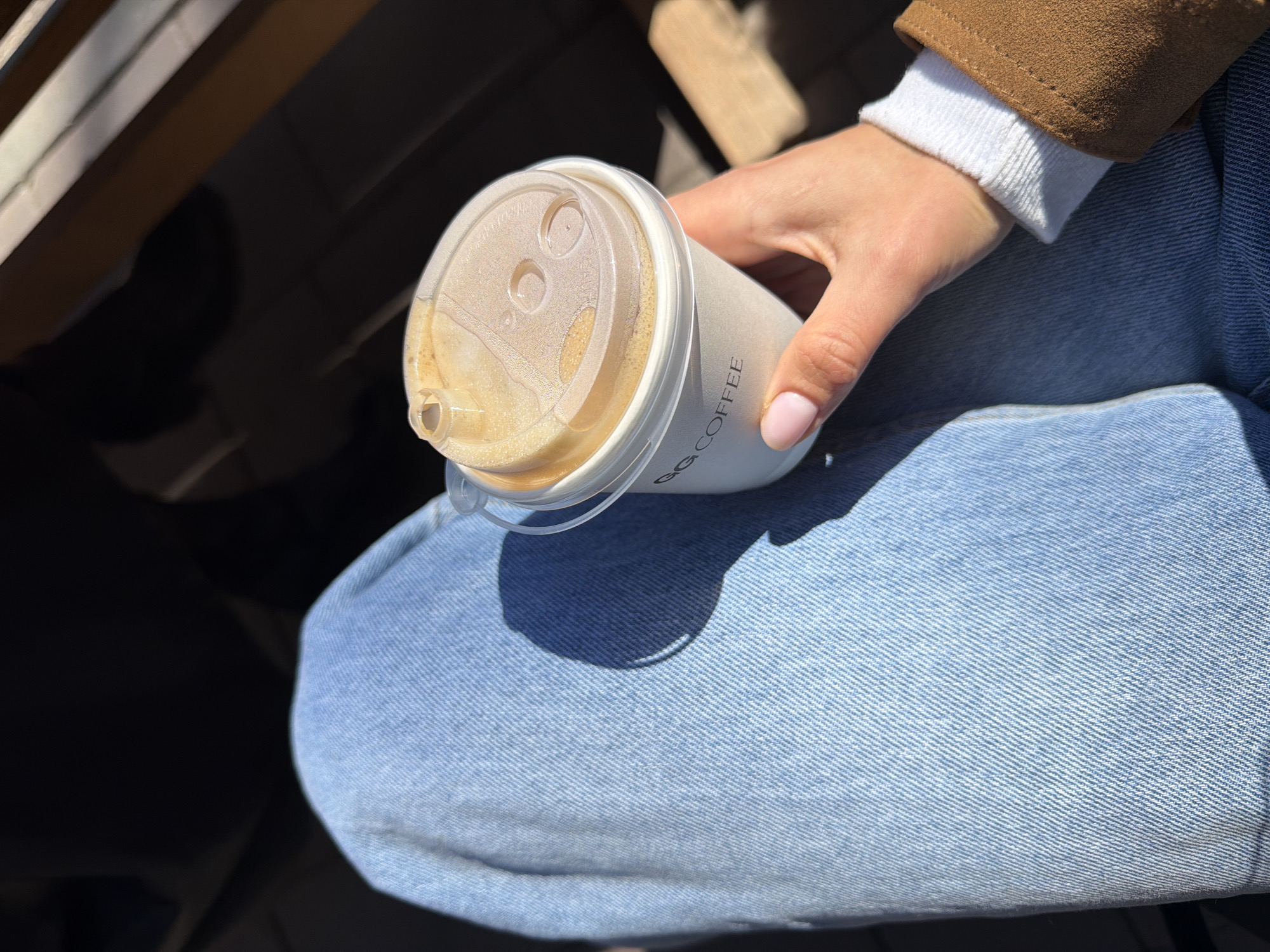A close-up, high-angle shot captures a person holding a coffee cup, likely taken outdoors or in a brightly lit indoor space in Krasnoyarsk, Russia. The individual is wearing light blue denim jeans, a white ribbed sweater or cuff, and a brown jacket. Their right hand, adorned with light pink nail polish on the thumb and index finger, grips a white takeaway coffee cup with a clear plastic lid. The cup contains a beverage with a frothy, light brown surface, typical of coffee or a similar drink.
Bright sunlight illuminates the scene, casting a distinct, dark circular shadow of the cup onto the person's lap, which is covered by the light blue jeans. The texture of the denim is clearly visible with its diagonal weave. In the blurred background, dark shadows and hints of wooden structures or furniture can be seen, suggesting the person might be seated at a table or bench. The bright lighting indicates it is daytime with clear weather.
Text visible on the side of the coffee cup reads "GG COFFEE" in a vertical orientation. The composition focuses on the hand, cup, and upper leg, with the rest of the environment largely out of focus, emphasizing the moment of enjoying a coffee.