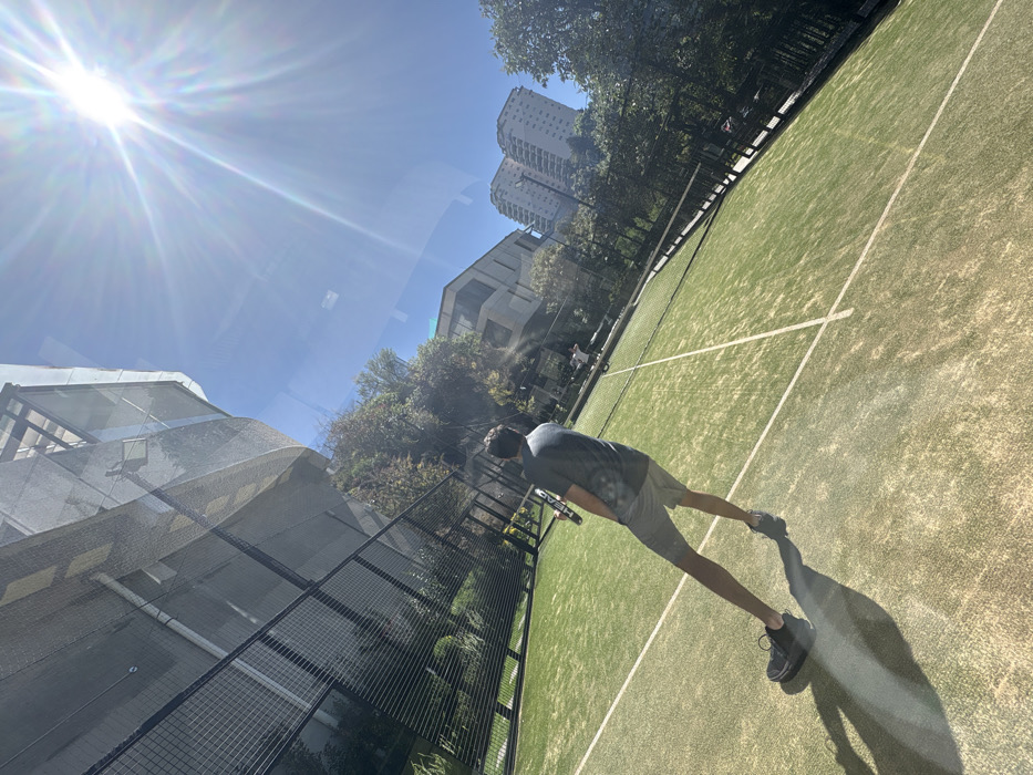 A man stands on a green sports court, holding a paddle racket, casting a long shadow due to the bright sun. A net bisects the court. Fencing surrounds the court perimeter. Trees and buildings are visible in the background, including a tall apartment building. The sun shines intensely in the upper left. The image appears to be taken through a reflective surface, creating a slightly distorted view.