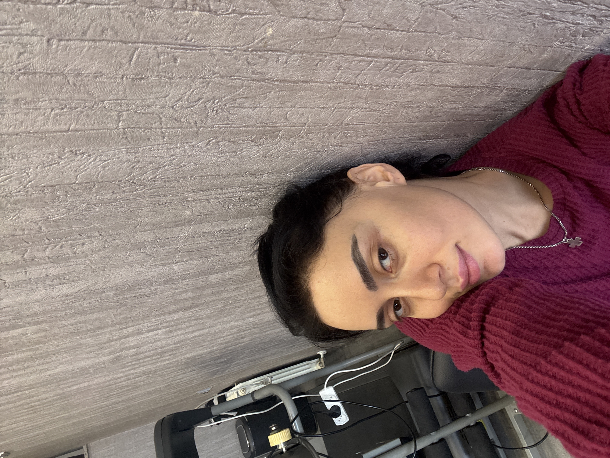 The scene depicts an adult female in an indoor setting, assumed to be in Tbilisi, Georgia. The woman, appearing to be in her late 20s to early 40s, has dark hair pulled back and brown eyes, looking directly towards the viewer with a slight smile. She wears a deep red, ribbed long-sleeved sweater and a silver-colored chain with a small cross pendant around her neck. Her right side rests against a light grey wall featuring a vertical, textured pattern. Her right arm, bent at the elbow, supports her head on her shoulder. In the upper left background, exercise equipment, including portions of a grey metal frame, black components, and yellow accents, is visible, suggestive of a treadmill or similar machine. A white power strip with multiple white electrical cords is plugged into it, lying on a surface below the equipment. A white panel radiator is partially visible behind the exercise equipment. The lighting is uniform, without strong directional shadows.
