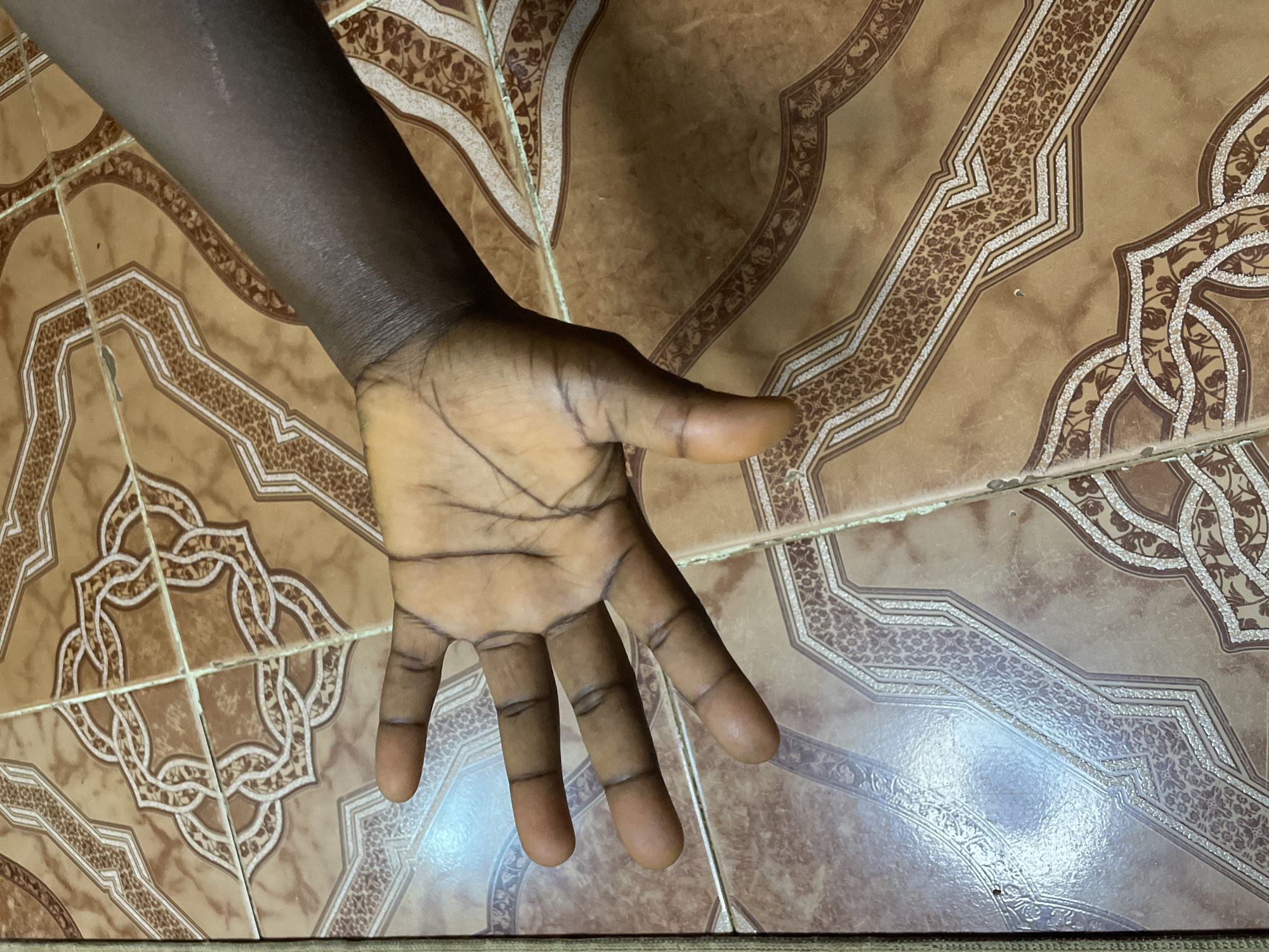 A close-up, high-angle shot shows a dark-skinned person's left hand and forearm resting on a tiled floor. The hand is open, palm facing upward, with the fingers slightly spread. The palm exhibits several prominent lines, and the skin appears dry. The forearm is also visible, showing a scar on the outer side. The floor is made of square tiles with a decorative pattern in shades of brown and beige, featuring geometric and floral motifs. The lighting is overhead, creating some glare on the tiles. The image was taken at an unknown location in the city of Narayi, Nigeria.