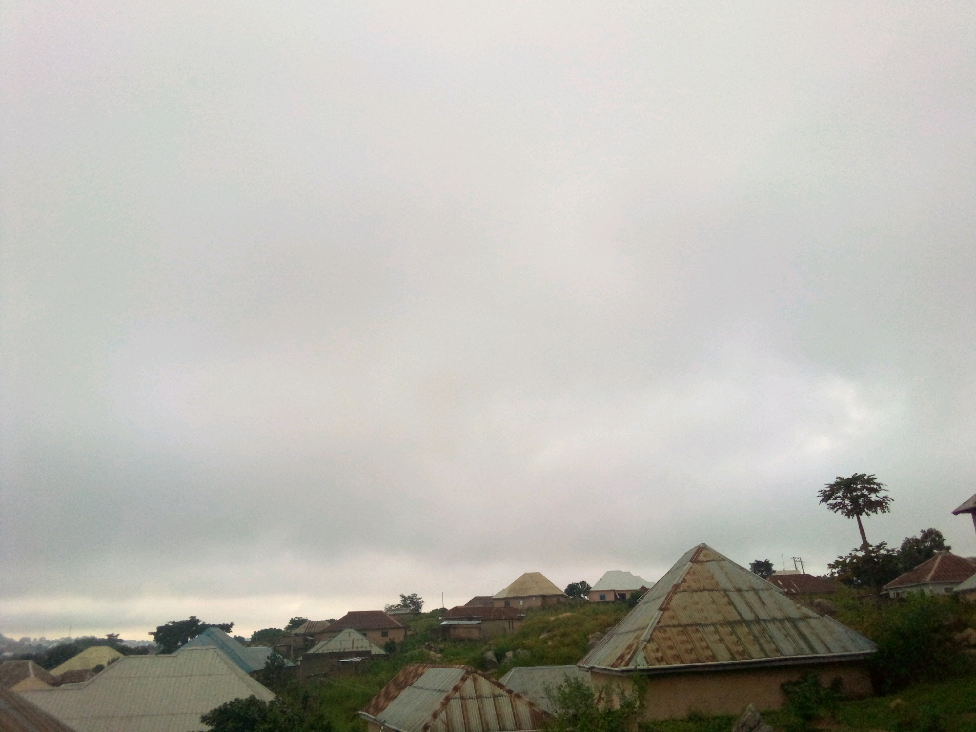 The image depicts a hillside settlement in Jos, Nigeria, under a heavily overcast sky. The foreground features several structures with distinctively angular, corrugated metal roofs, some showing signs of age and rust. These buildings are clustered on the slope, with some appearing more complete than others. In the middle ground and background, more houses are visible, also nestled into the terrain. Some have peaked roofs, while others have flatter, more irregular shapes. Vegetation, including trees and bushes, is interspersed among the buildings, indicating a semi-rural or suburban setting. A prominent, tall, slender tree with a sparse canopy stands out against the sky on the right side of the image. The overall atmosphere is muted due to the cloudy weather. There are no people visible in the image, nor any signs of vehicles or significant activity. The scene is a static view of residential architecture integrated with the natural landscape of the hilly terrain.