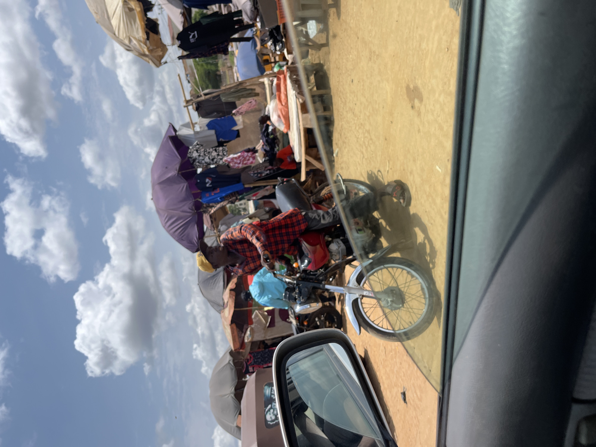 The image, taken from inside a vehicle, shows a street scene in Galadimawa Village, Nigeria. A man on a motorcycle is positioned in the street near roadside vendors. The vendors are displaying clothing for sale, with some items hanging under umbrellas for shade. The sky is visible in the background and features scattered clouds. The vehicle's side mirror and window frame are visible in the foreground.