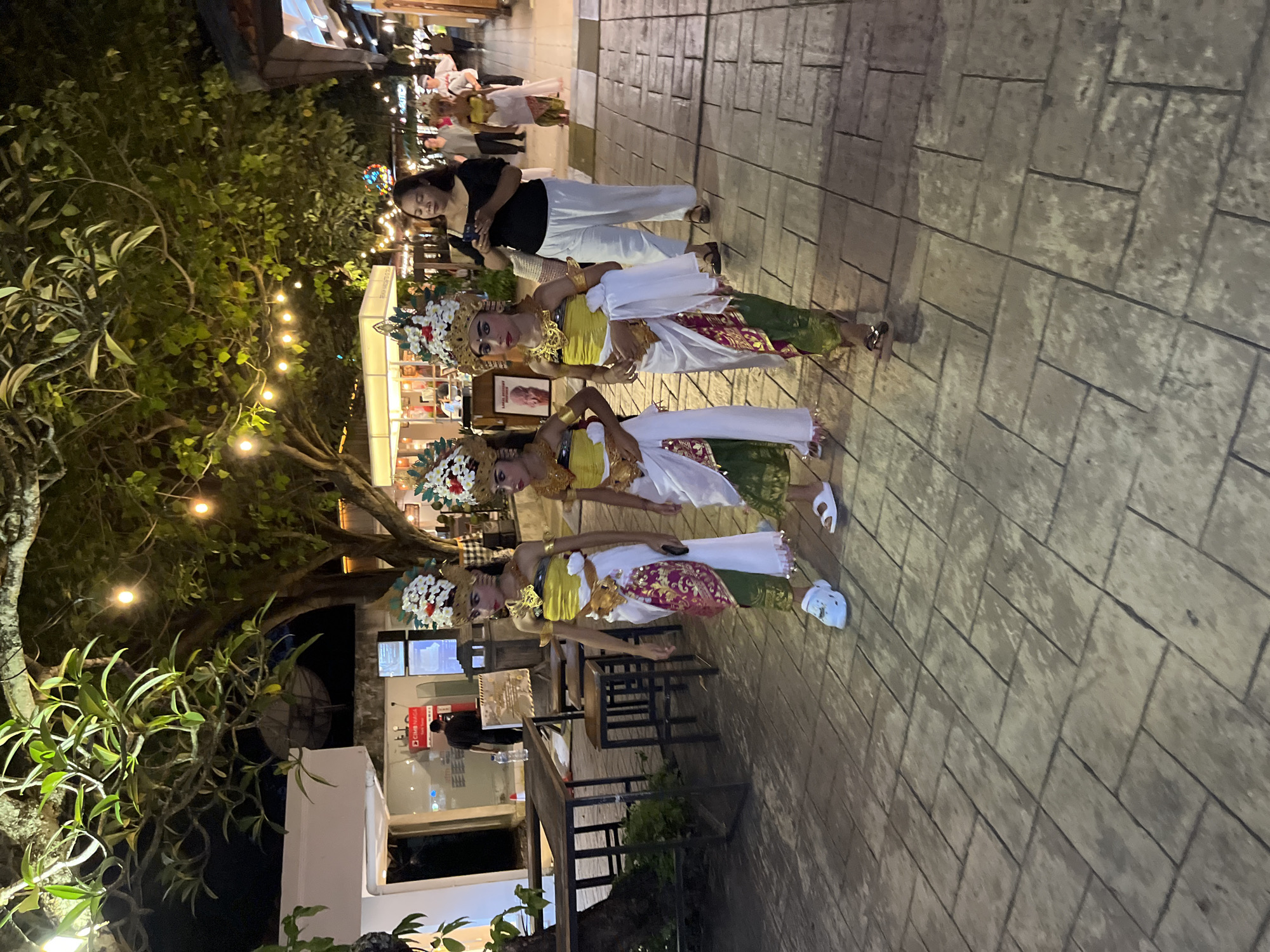 At night in Kuta Selatan, Indonesia, three women in traditional Balinese dance attire pose on a paved walkway. A woman in a black top and white pants is taking their picture. Nearby, trees with string lights hang overhead, and tables and chairs are set up outside a building.