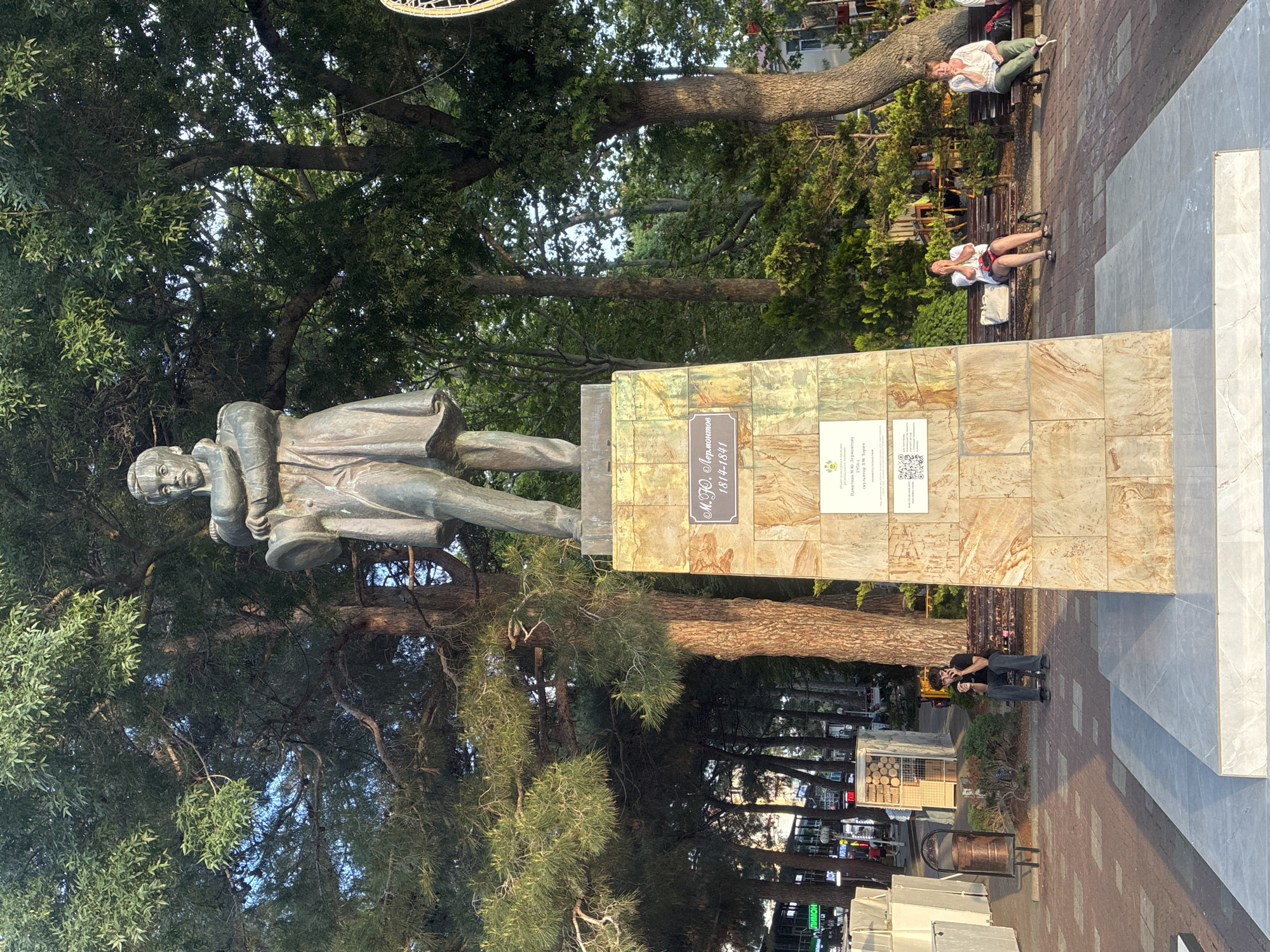 In the city of Gelendzhik, Russia, a bronze statue of Mikhail Lermontov stands on a stone pedestal adorned with inscriptions. The statue depicts Lermontov in military attire, holding a hat. Behind the statue, lush green trees create a natural backdrop. In the foreground, a paved walkway surrounds the monument, and several people are visible. A young man sits on a bench, looking at his phone. Further back, two women sit on benches, one appearing to be covering her face. The overall scene suggests a public space, possibly a park or promenade, during daylight hours.
